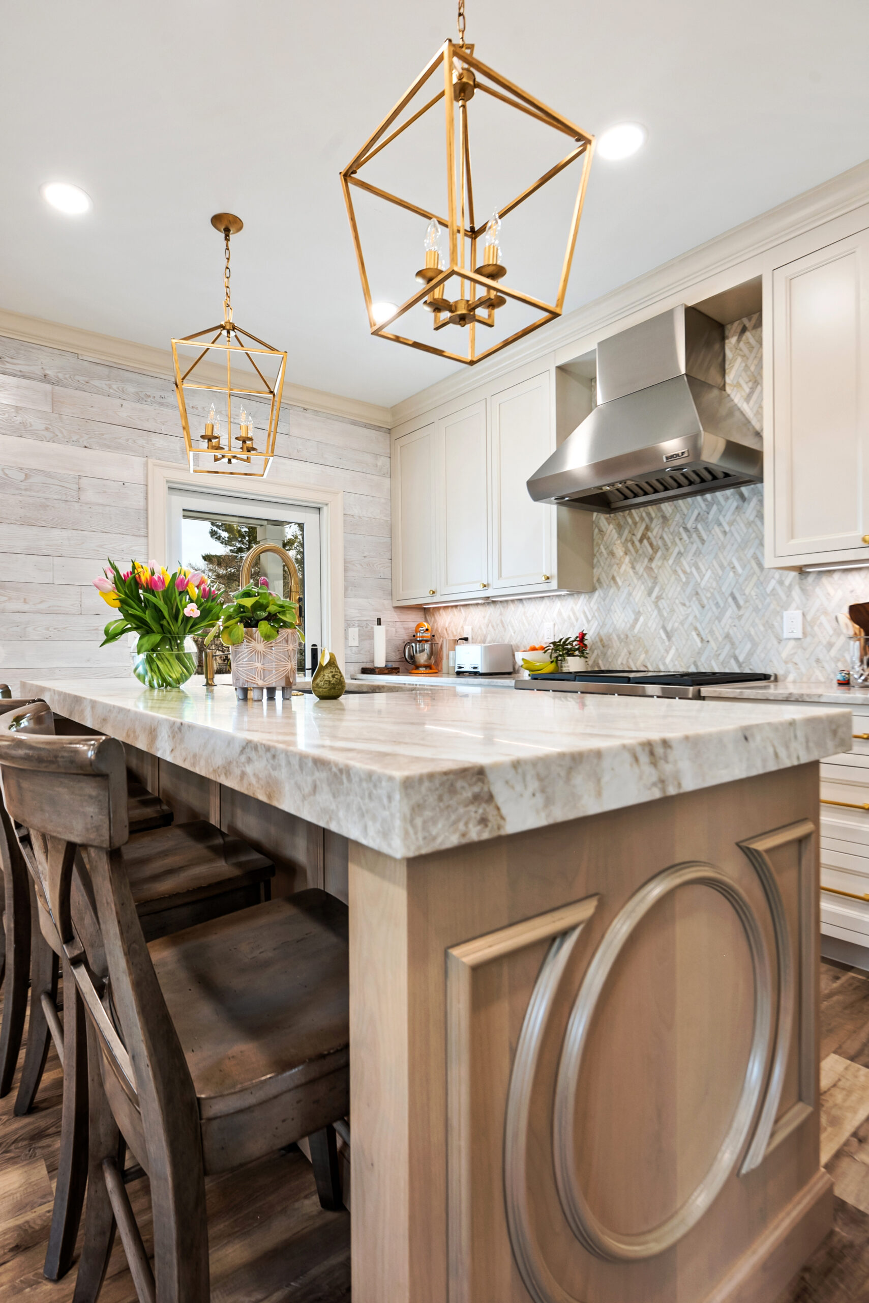Modern kitchen remodel with a large island, wooden chairs, marble countertop, gold pendant lights, stainless steel range hood, and decorative flowers. Light wood cabinets and backsplash complete the space by a top kitchen remodeling company in Columbus, Ohio.