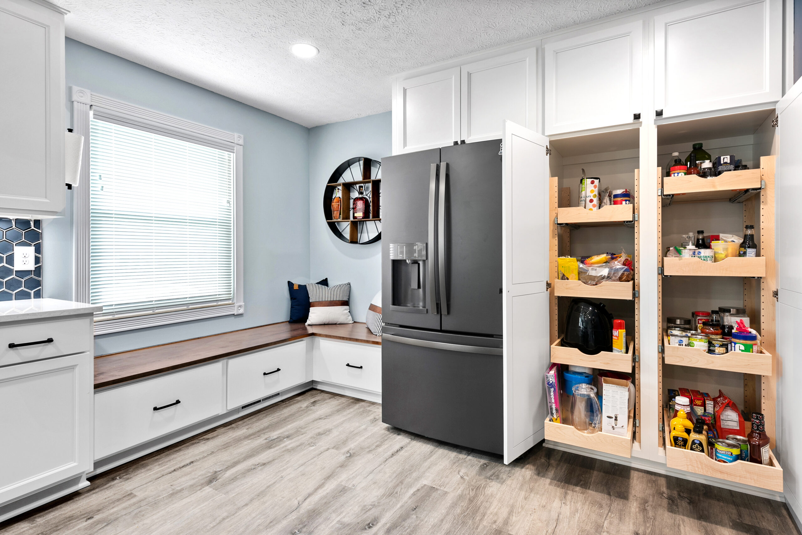 Modern kitchen remodel with light blue walls, a large refrigerator, an open pantry stocked with food, white cabinets, wood flooring, a cozy window seat with cushions, and a round black wall shelf.