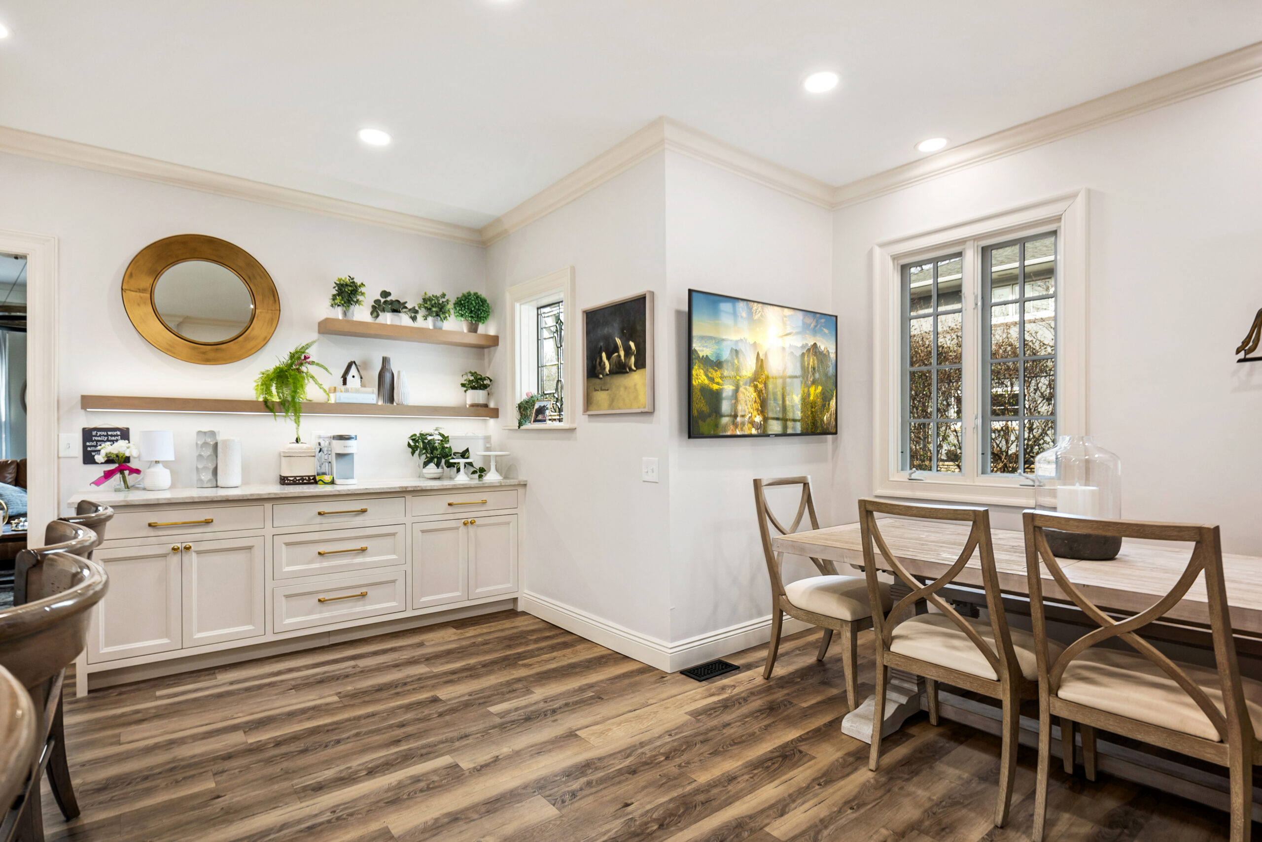 Bright kitchen and dining area with white cabinets, open shelves, potted plants, a round mirror, wooden dining table with four chairs, large wall art, and wood-patterned flooring under recessed lighting—perfect inspiration for your next bath remodel.