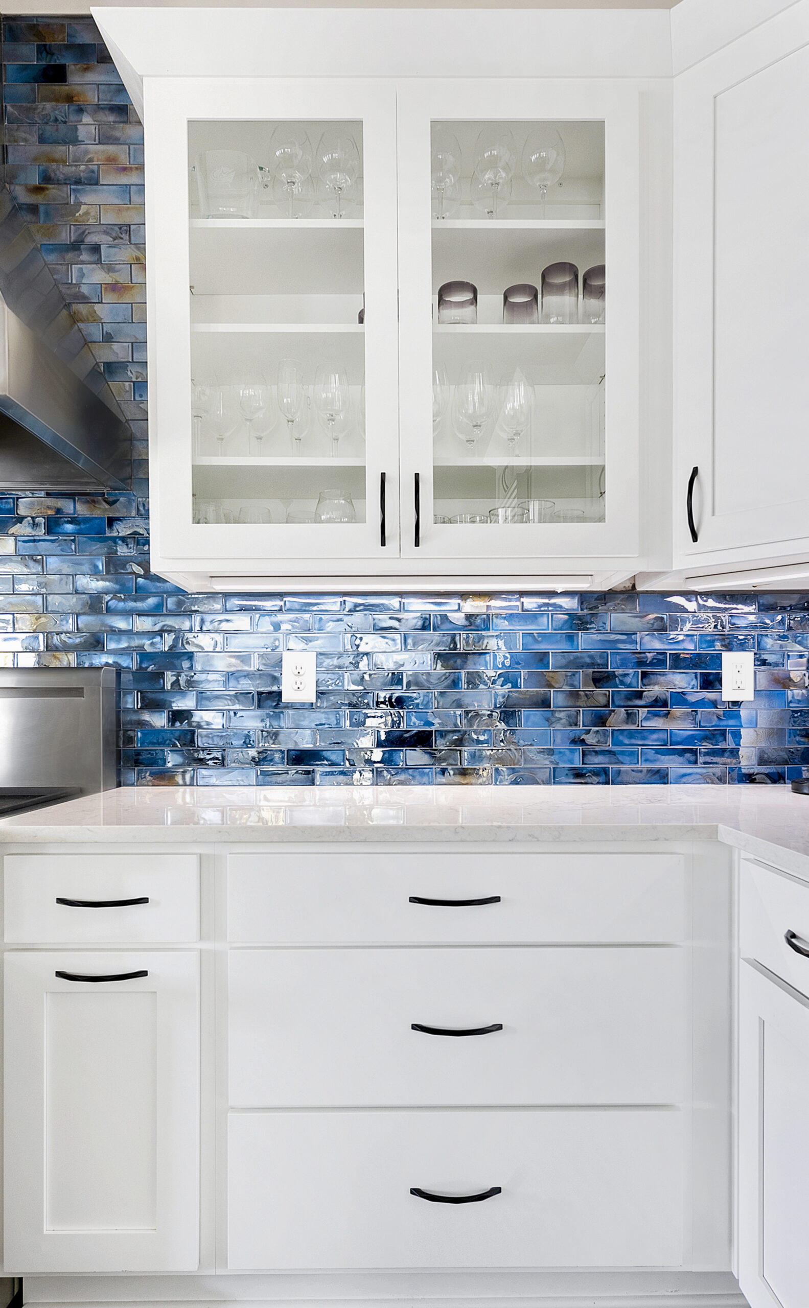 A modern kitchen remodel with white cabinets and drawers, glass-front upper cupboards displaying glassware, a glossy blue tiled backsplash, white countertops, and sleek black handles on the cabinetry.