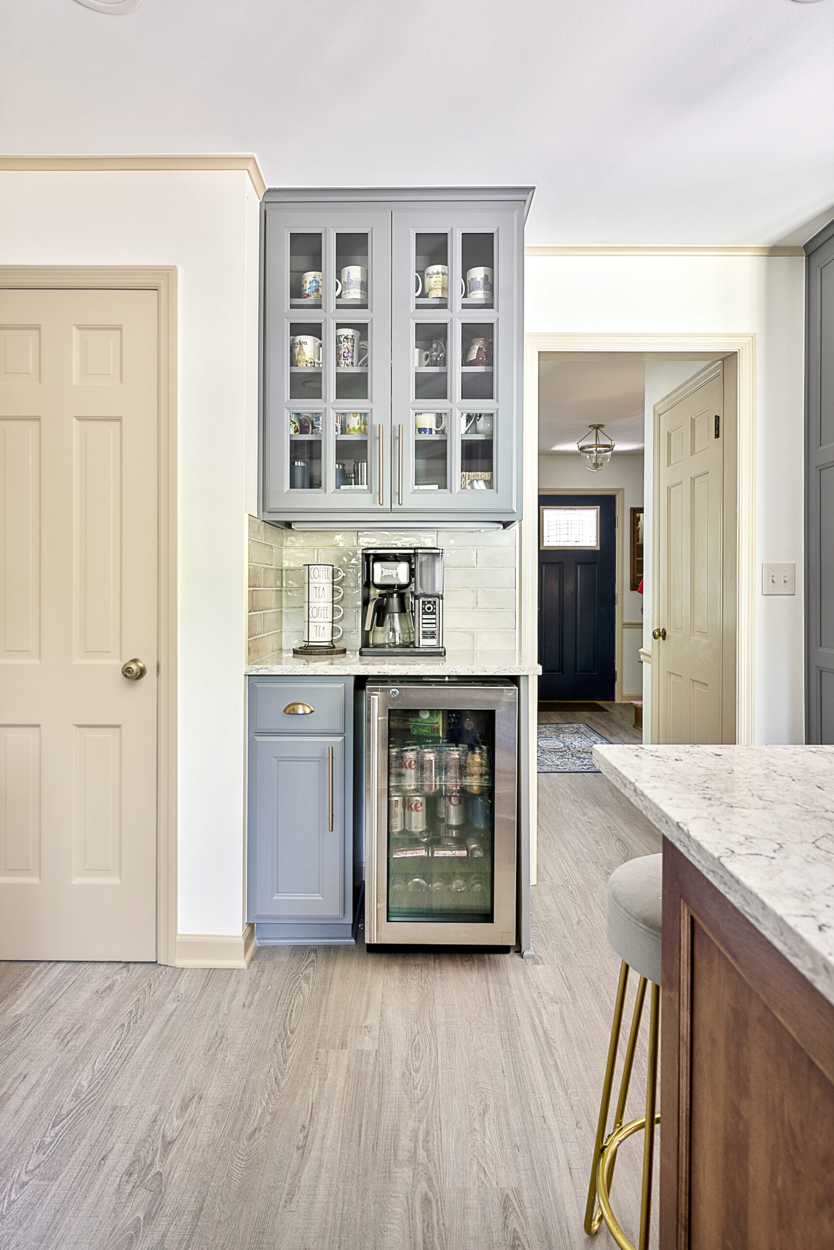 A small home coffee bar with light blue cabinets, a built-in mini fridge stocked with drinks, a coffee maker, glass-front upper cabinets, and light wood flooring—perfect inspiration from a kitchen remodel by a top kitchen remodeling company Columbus Ohio.