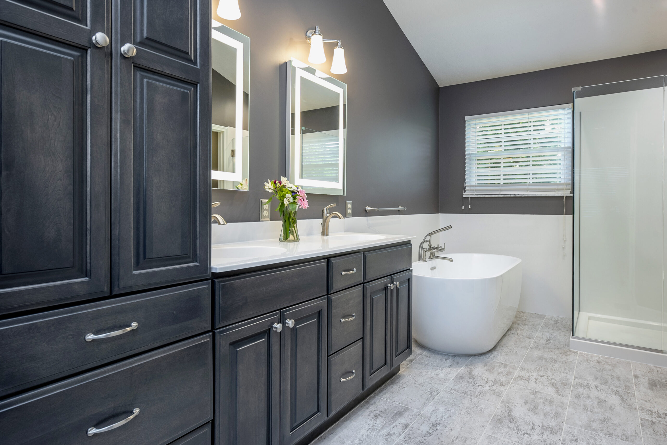 Modern bathroom with dark wood cabinets, double sinks with mirrors, a white freestanding bathtub, a glass shower, and gray tile floor. Perfect for your next bath remodel, this space features a window with blinds and flowers on the counter.