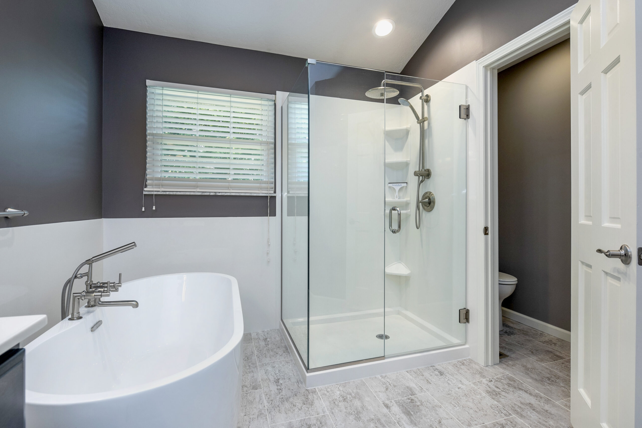 Modern bathroom with a freestanding white bathtub, glass-enclosed shower, gray walls, and large window with blinds. Designed by a top kitchen remodeling company Columbus Ohio. Light gray tile flooring leads to a private toilet area.