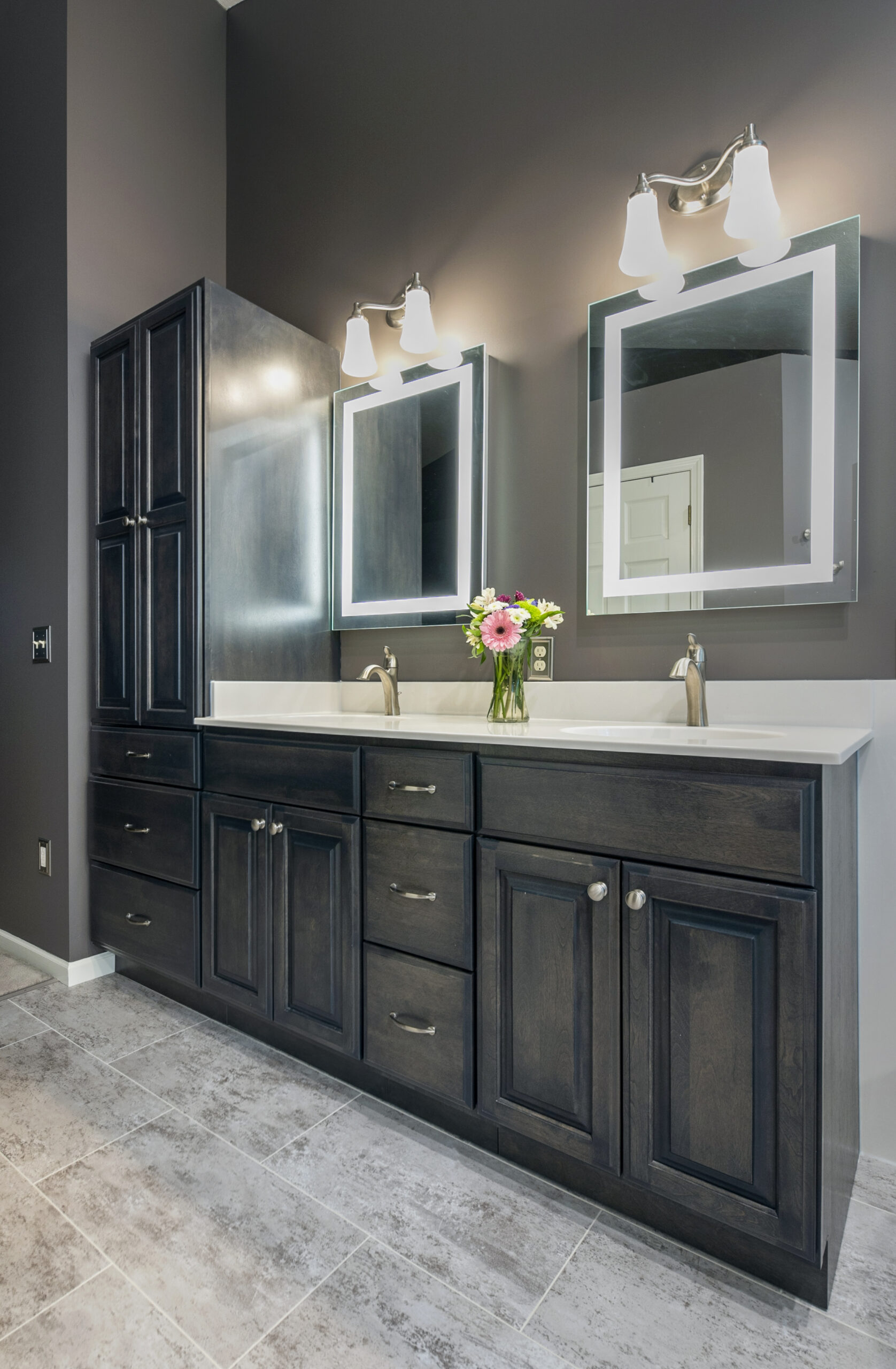 Modern bathroom renovation featuring a dark wood double vanity, two rectangular mirrors with lights above each sink, a vase of flowers on the counter, gray tiled floor, and neutral-toned walls.
