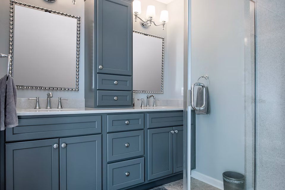 Modern bathroom with blue-gray cabinets, double sinks, chrome fixtures, wall lights, a towel ring, and a glass shower door. Expertly designed by a leading kitchen remodeling company Columbus Ohio. Light walls and countertops brighten the space.