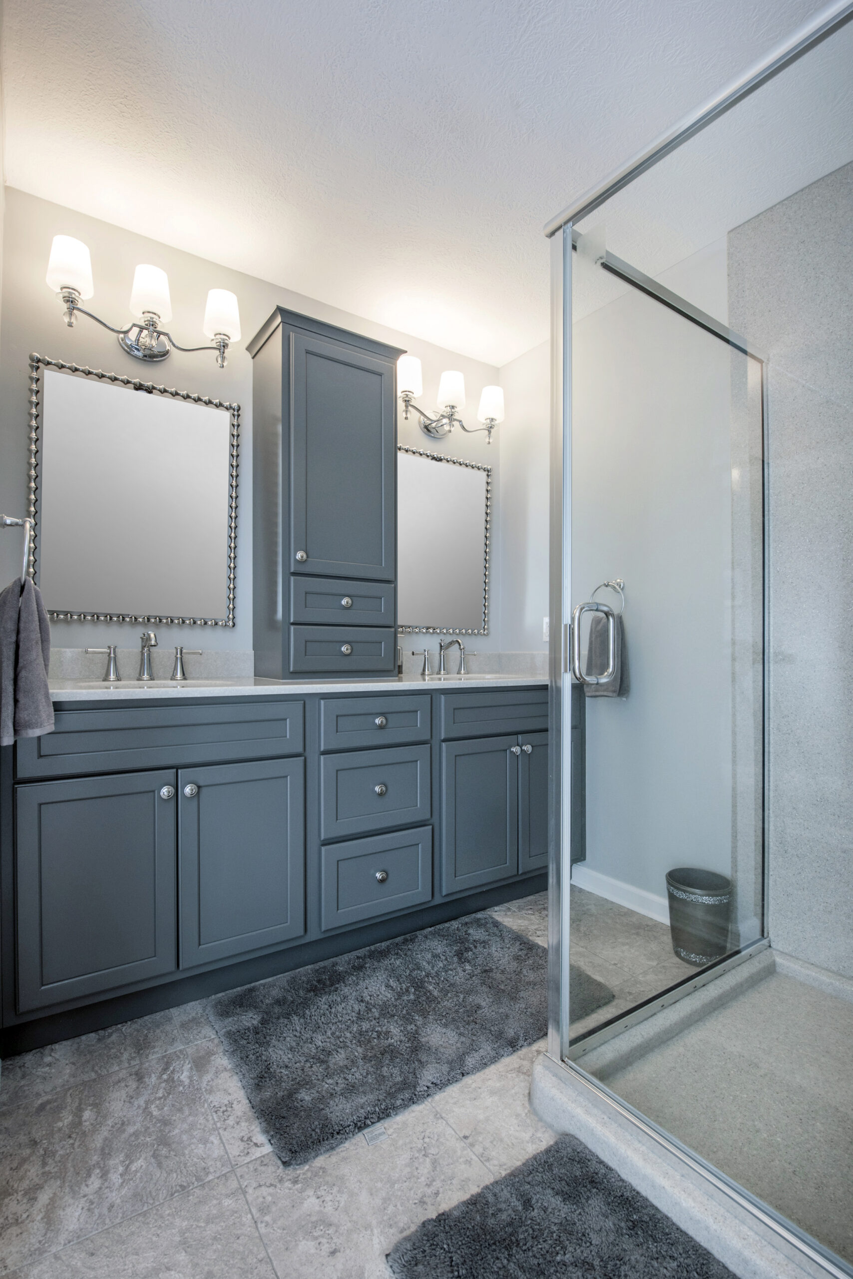 Modern bathroom renovation with gray cabinetry, two mirrors, double sinks, wall sconces above each mirror, glass shower enclosure, marble tile floor, and gray bath mats. A small trash bin is visible inside the shower area.