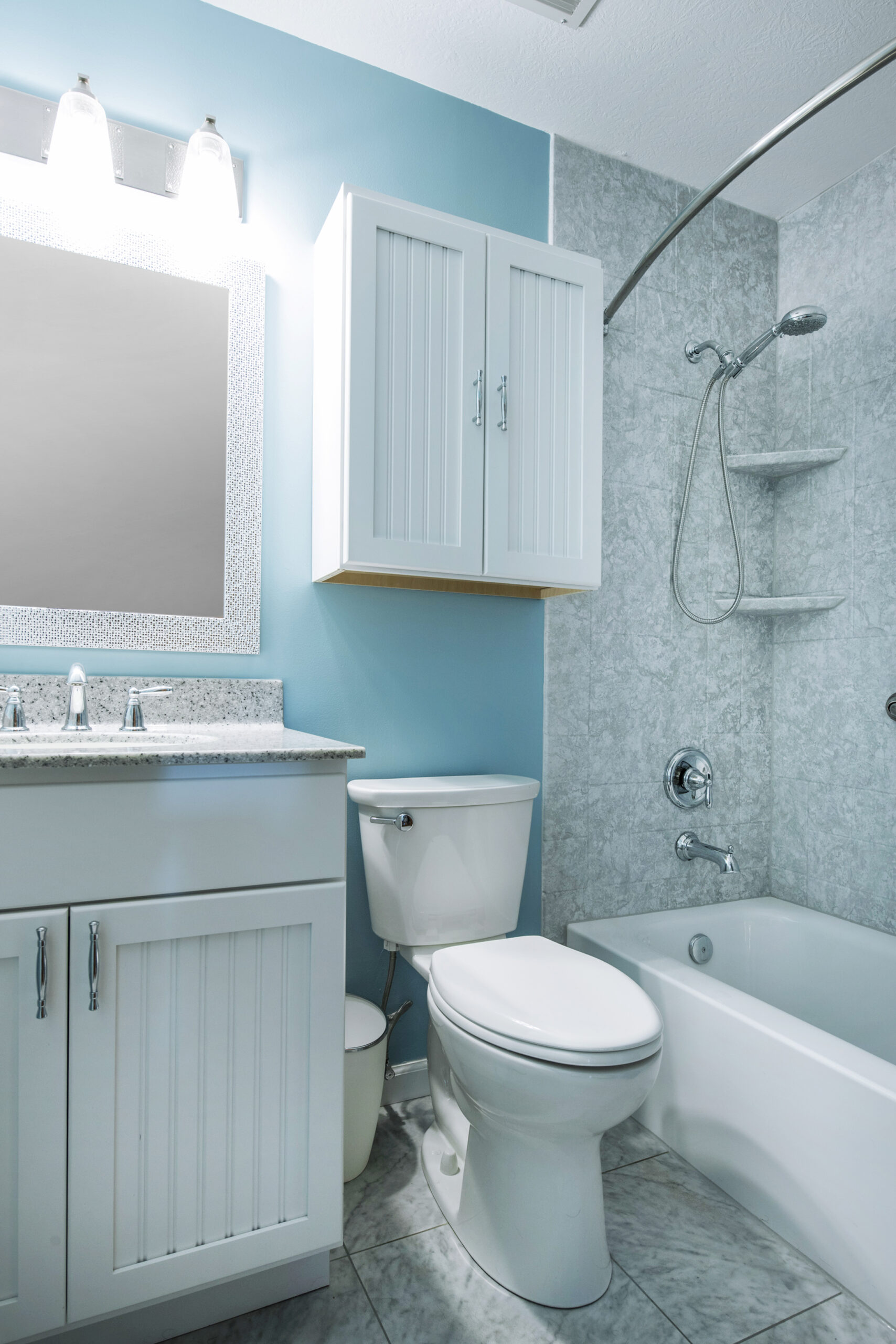Modern bathroom with white cabinetry, a toilet, and a bathtub with shower. Light blue and gray walls, large mirror above the sink, overhead lighting, and marble-like tile flooring—perfect inspiration for your bathroom renovation.