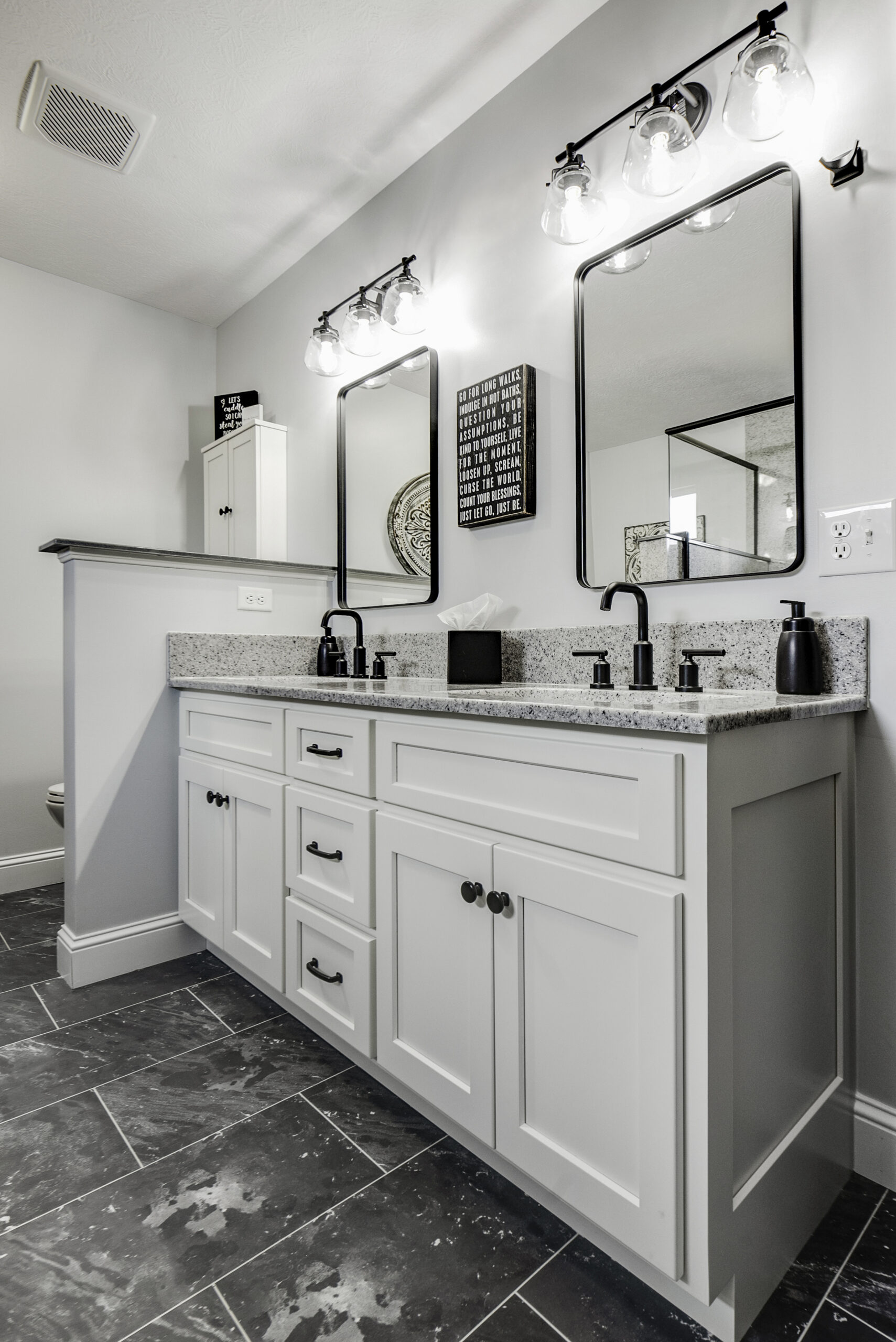 Modern bathroom by a leading bathroom remodeling company in Columbus, Ohio, featuring double white vanity, granite countertop, two black framed mirrors, black fixtures, wall lights above each mirror, and dark gray marble floor tiles with elegant decor.