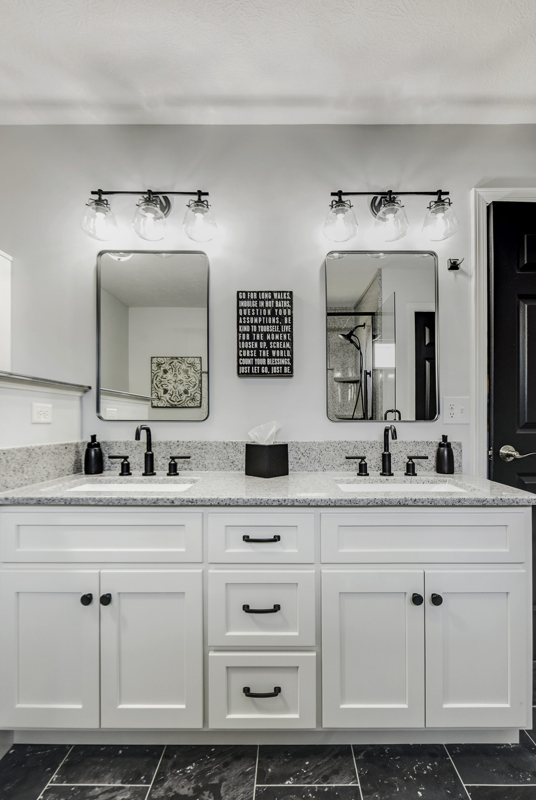 Modern bathroom with double sink vanity, granite countertop, black faucets and hardware, black soap dispensers, tissue box, and decorative wall art between the mirrors—a perfect example of a stylish bath remodel. Marble tile floor completes the look.