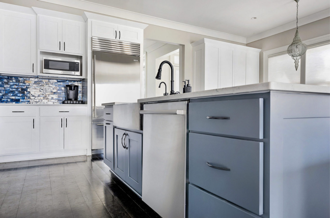 Modern kitchen renovation with white upper cabinets, blue lower island cabinets, stainless steel appliances, a farmhouse sink, and a blue mosaic tile backsplash. Dark wood flooring contrasts with the light cabinetry.