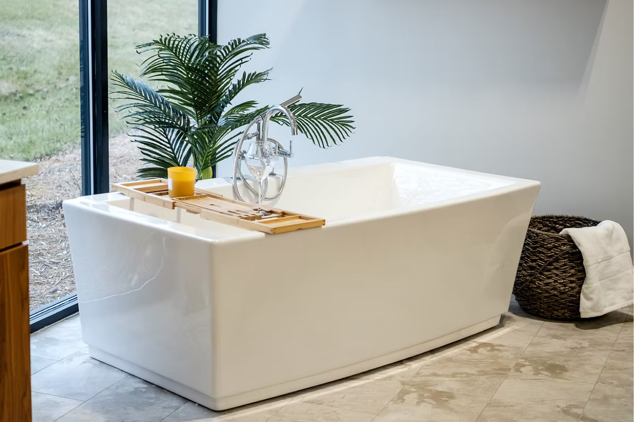 A modern white bathtub sits on a tiled floor near a large window, with a wooden tray holding a yellow candle and a plant beside it—an inviting space created by a leading bathroom remodeling company in Columbus, Ohio. A woven basket with a white towel is nearby.