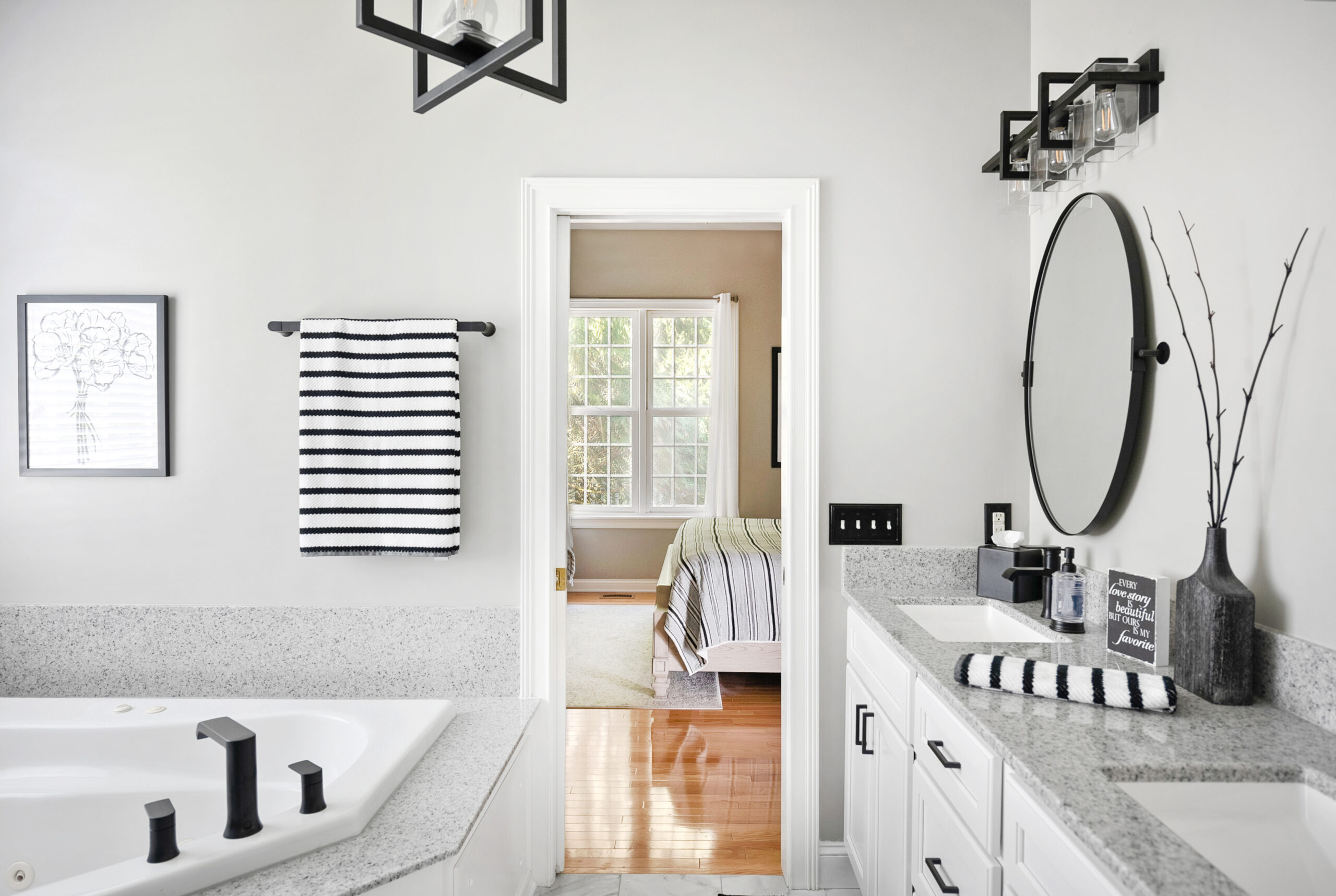 Modern bathroom with white cabinets, a granite countertop, black fixtures, and striped black-and-white towels—a perfect example of a stylish bath remodel. A doorway leads to a bright bedroom with large windows and a neatly made bed.