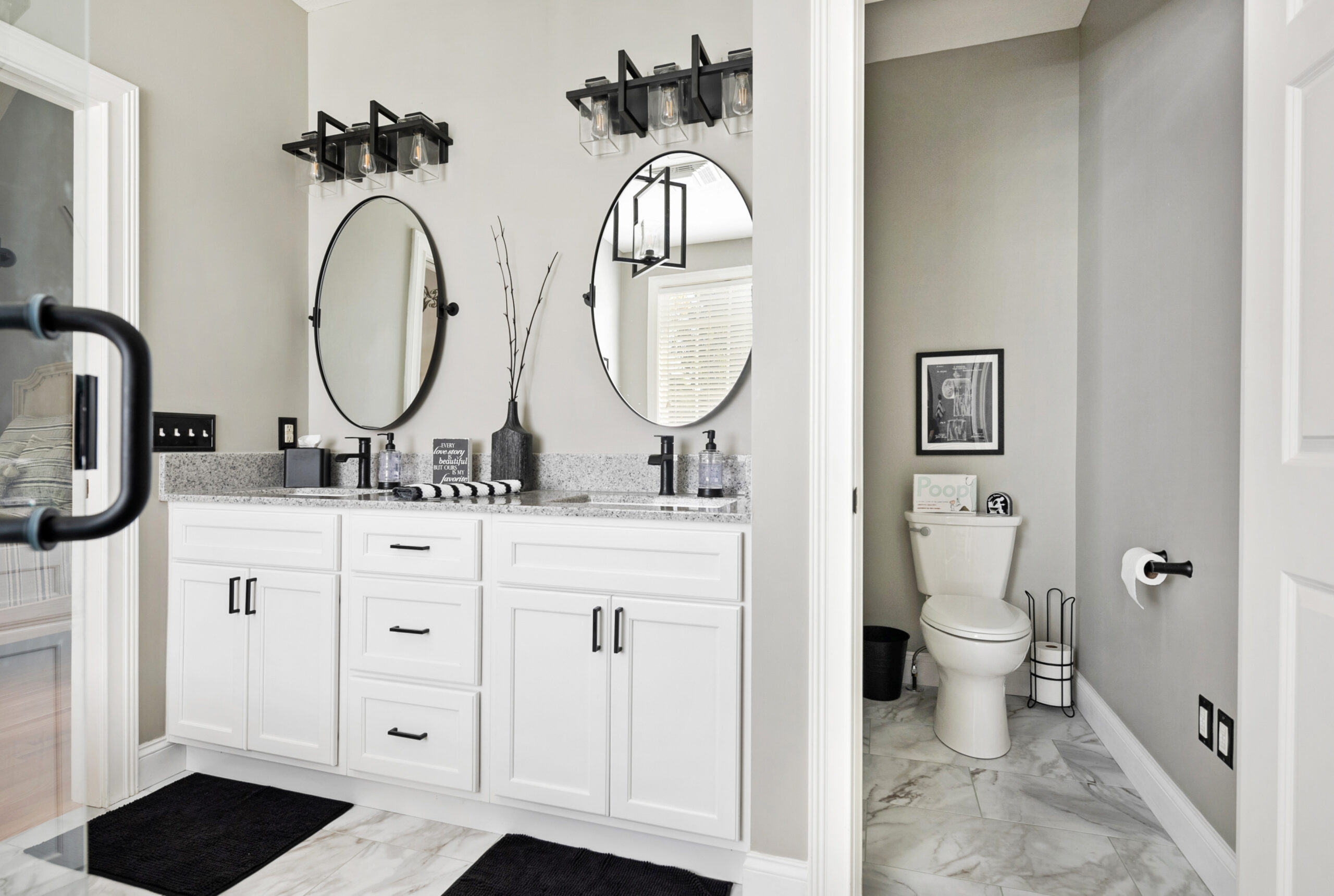 Modern bathroom with double vanity, round mirrors, and black fixtures. The countertop features decor and soap dispensers. Designed with the expertise of a kitchen remodeling company in Columbus, Ohio, the look continues into the adjoining room.