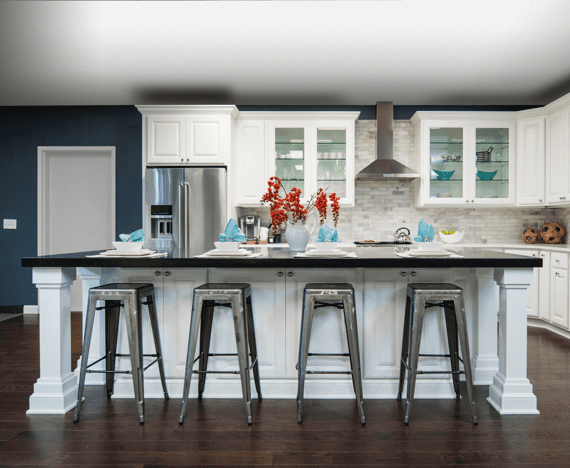 Modern kitchen renovation with white cabinets, a large island with four metal stools, stainless steel appliances, glass-front cabinets, dark countertops, and a vase with red flowers on the island.