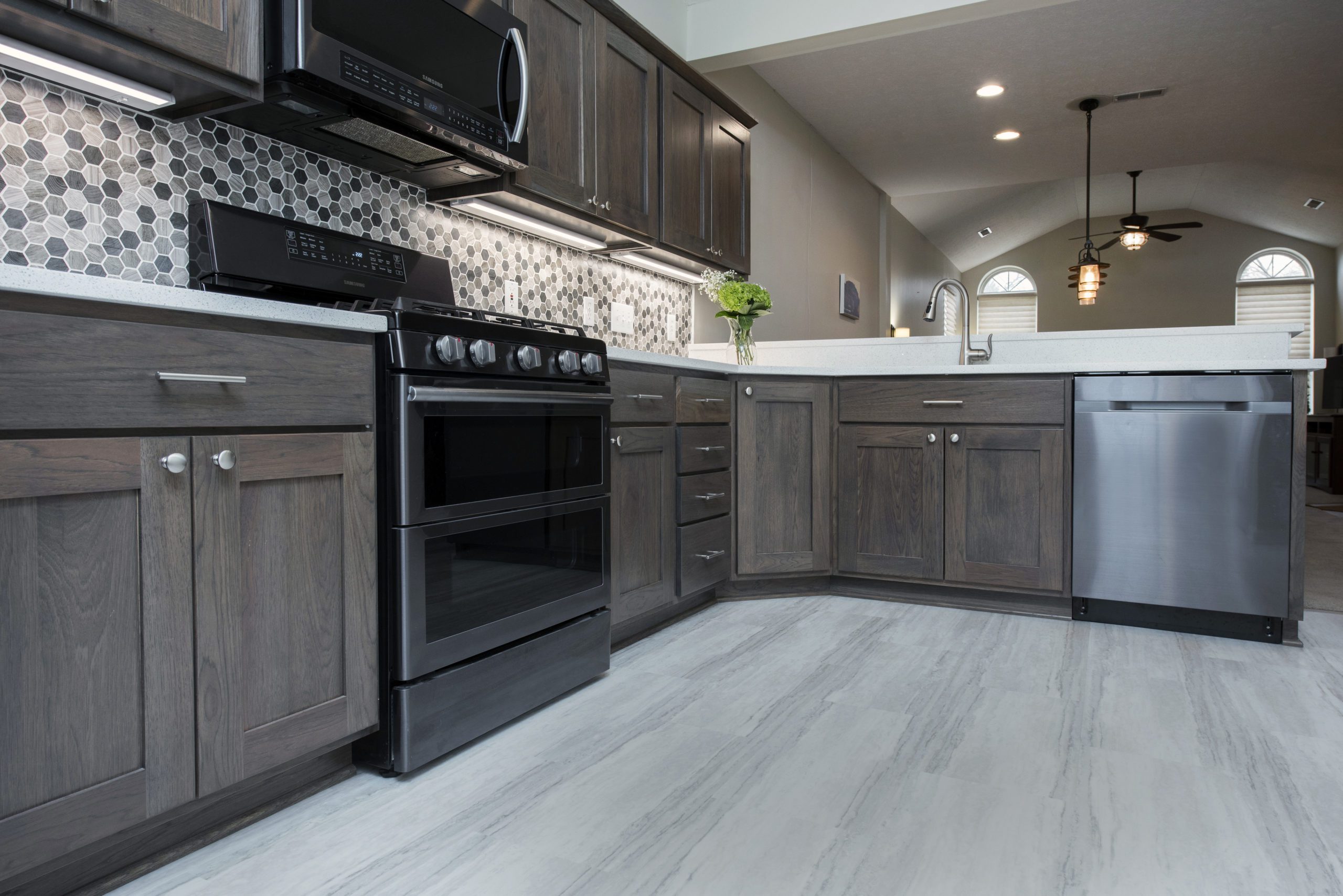 Modern kitchen remodel with dark wooden cabinets, black appliances, hexagon tile backsplash, light countertops, and grey flooring. A vase with green flowers sits near the sink—designed by a leading kitchen remodeling company in Columbus, Ohio.