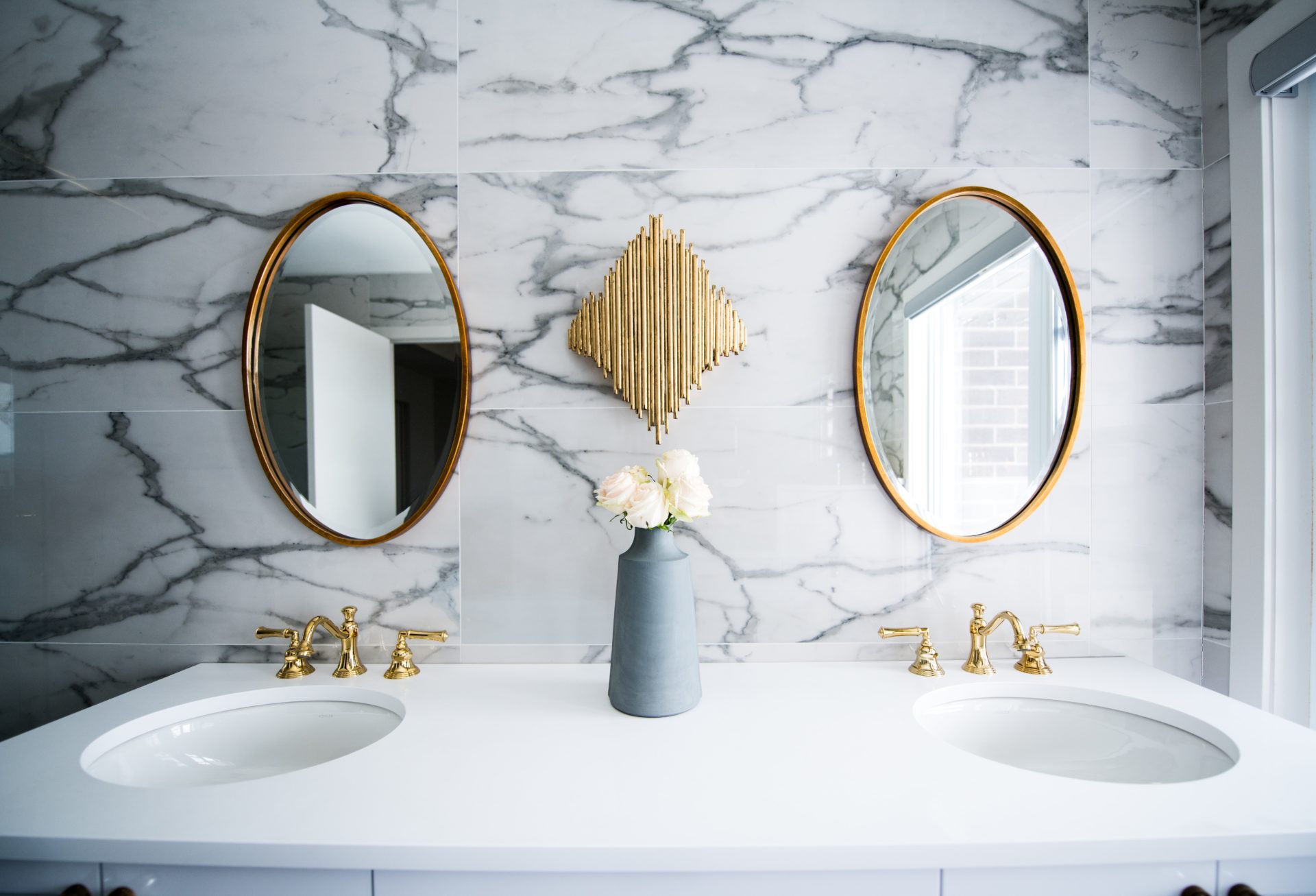 A modern bathroom renovation with a double sink, gold faucets, oval mirrors with gold frames, a gray vase holding white flowers, and marble-patterned wall tiles. A decorative gold wall accent is centered above the vase.