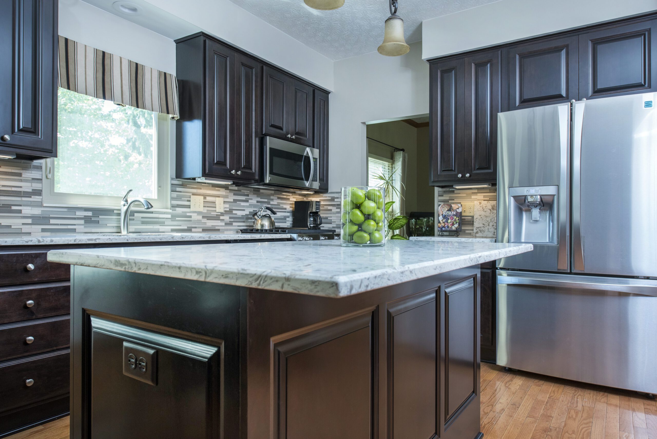Modern kitchen remodel with dark wood cabinets, stainless steel appliances, marble countertops, a center island, and a glass vase filled with green apples. Light streams in from a window with a striped valance.