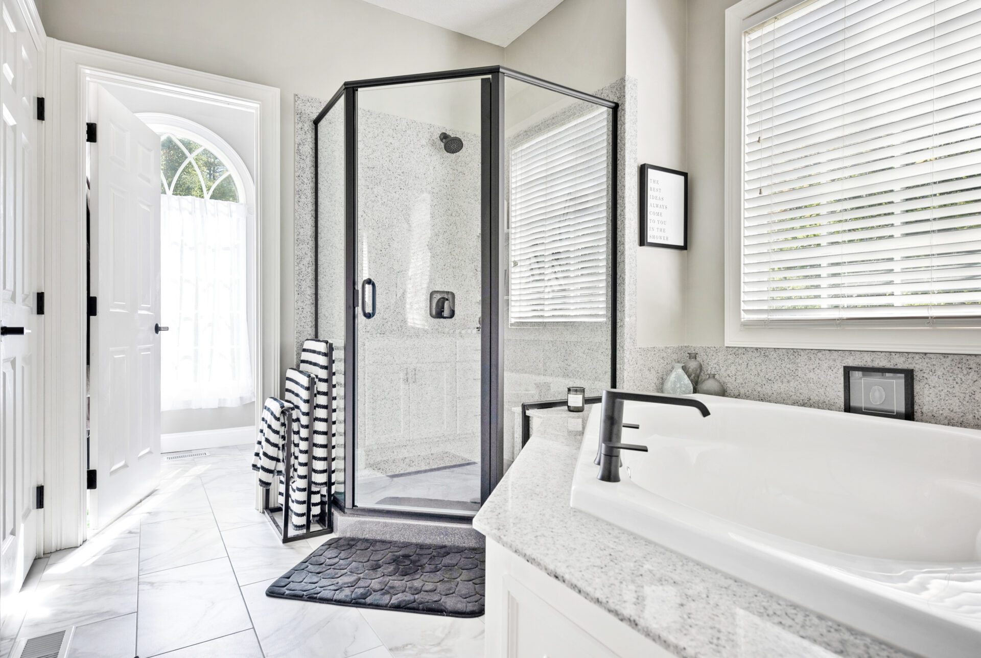 Bright, modern bathroom with a glass shower, white bathtub, black fixtures, striped towels stacked near the shower—beautifully updated by a leading bathroom remodeling company in Columbus, Ohio. White marble floors and countertops complete the look.