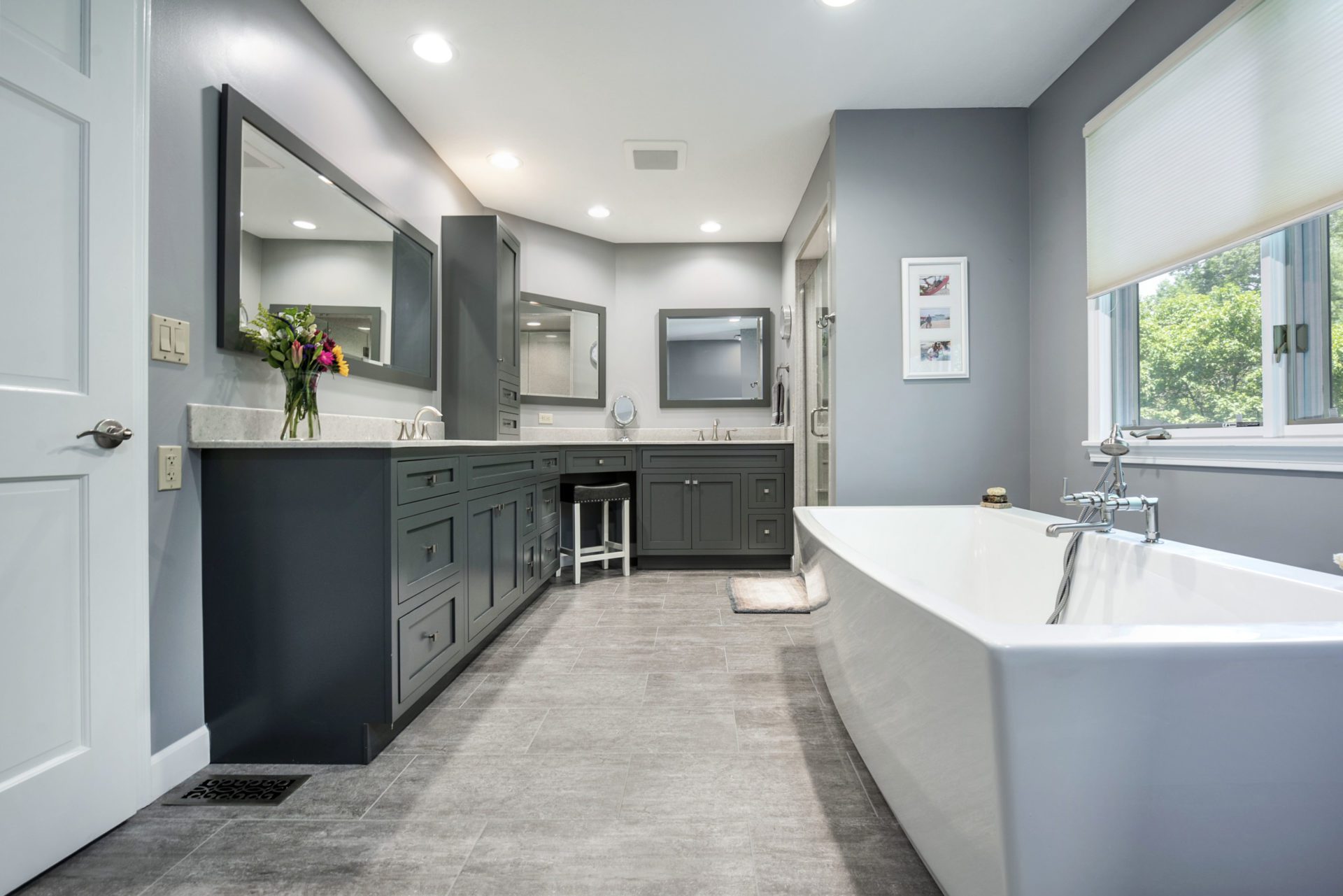 Spacious modern bathroom with gray cabinets, large mirrors, a freestanding white bathtub, and a window with a view of greenery—an inviting space crafted by a leading bathroom remodeling company Columbus Ohio. Elegant decor includes flowers on the vanity.