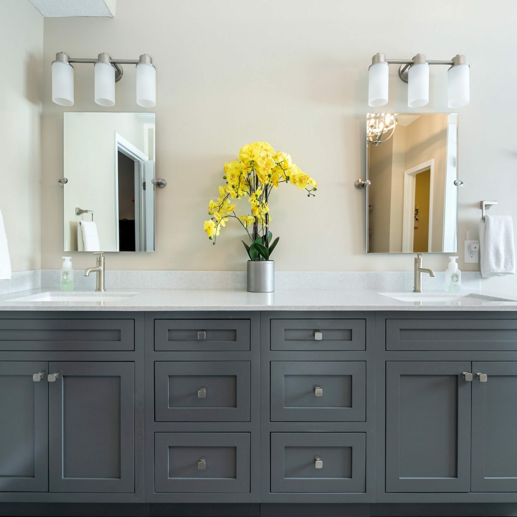 A modern bathroom renovation with a gray double-sink vanity, white countertop, two mirrors, wall-mounted lights, and a vase of bright yellow flowers in the center.