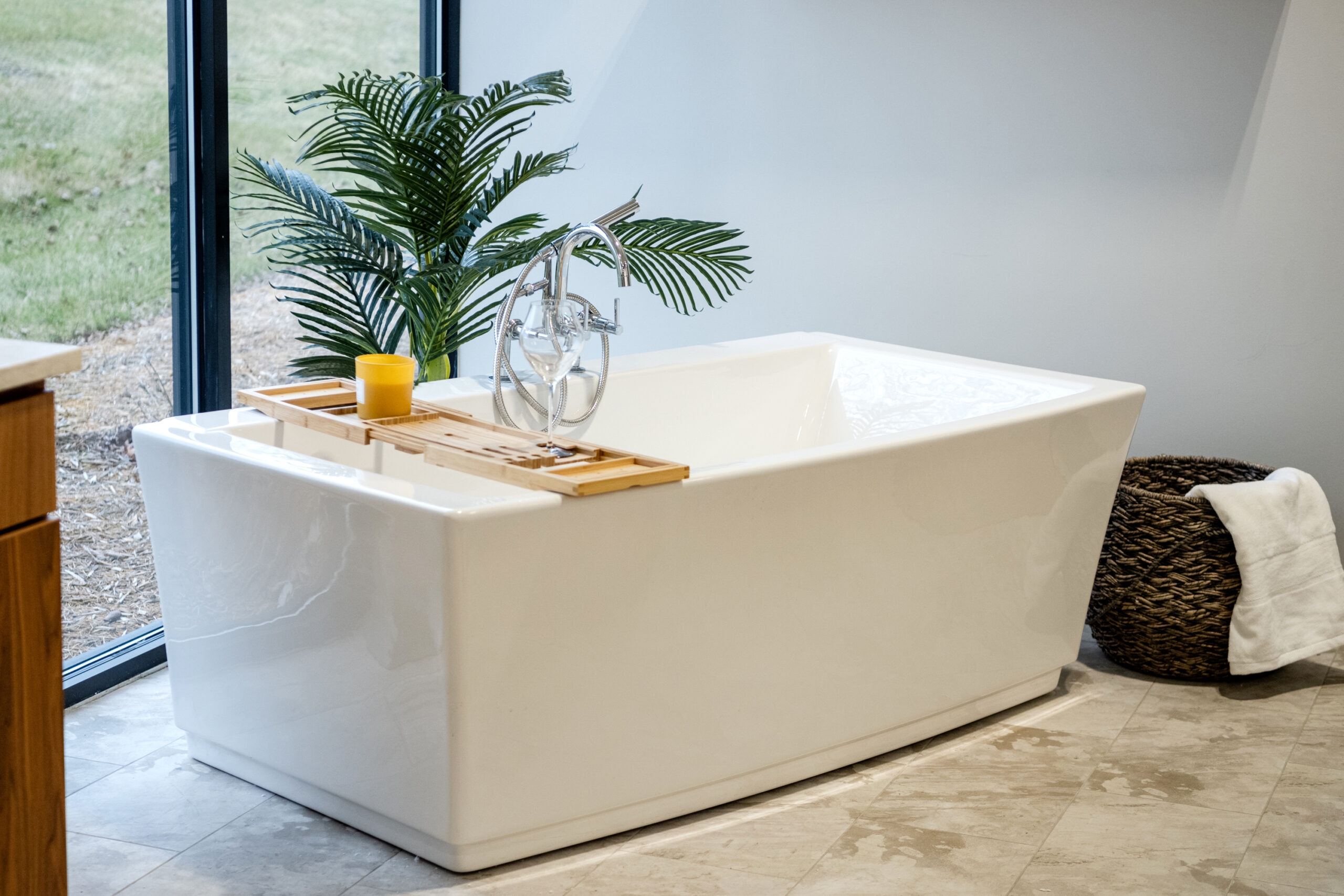 A modern white bathtub sits by a large window with a green plant and wooden caddy holding a candle and glass—perfect inspiration for your bath remodel from a leading bathroom remodeling company in Columbus, Ohio.