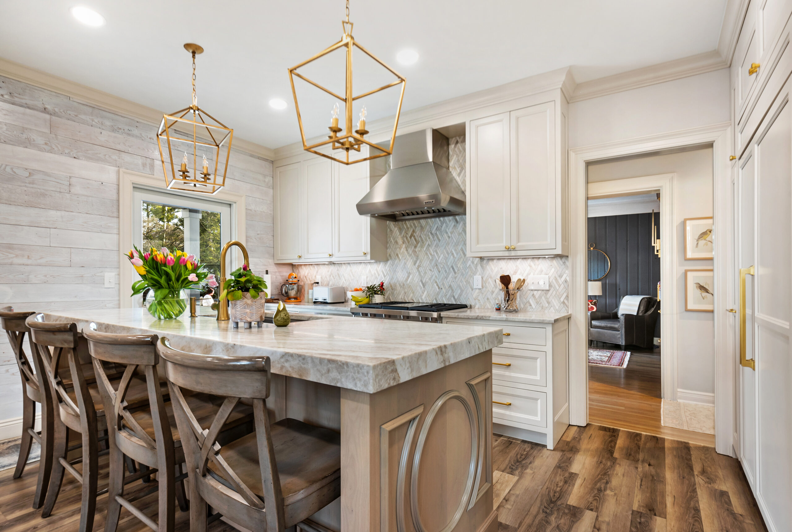 Modern kitchen with light wood cabinets, a large island with wooden chairs, two gold pendant lights, fresh flowers on the countertop, stainless steel appliances, and wood flooring—crafted by a top kitchen remodeling company Columbus Ohio. Living room visible through doorway.