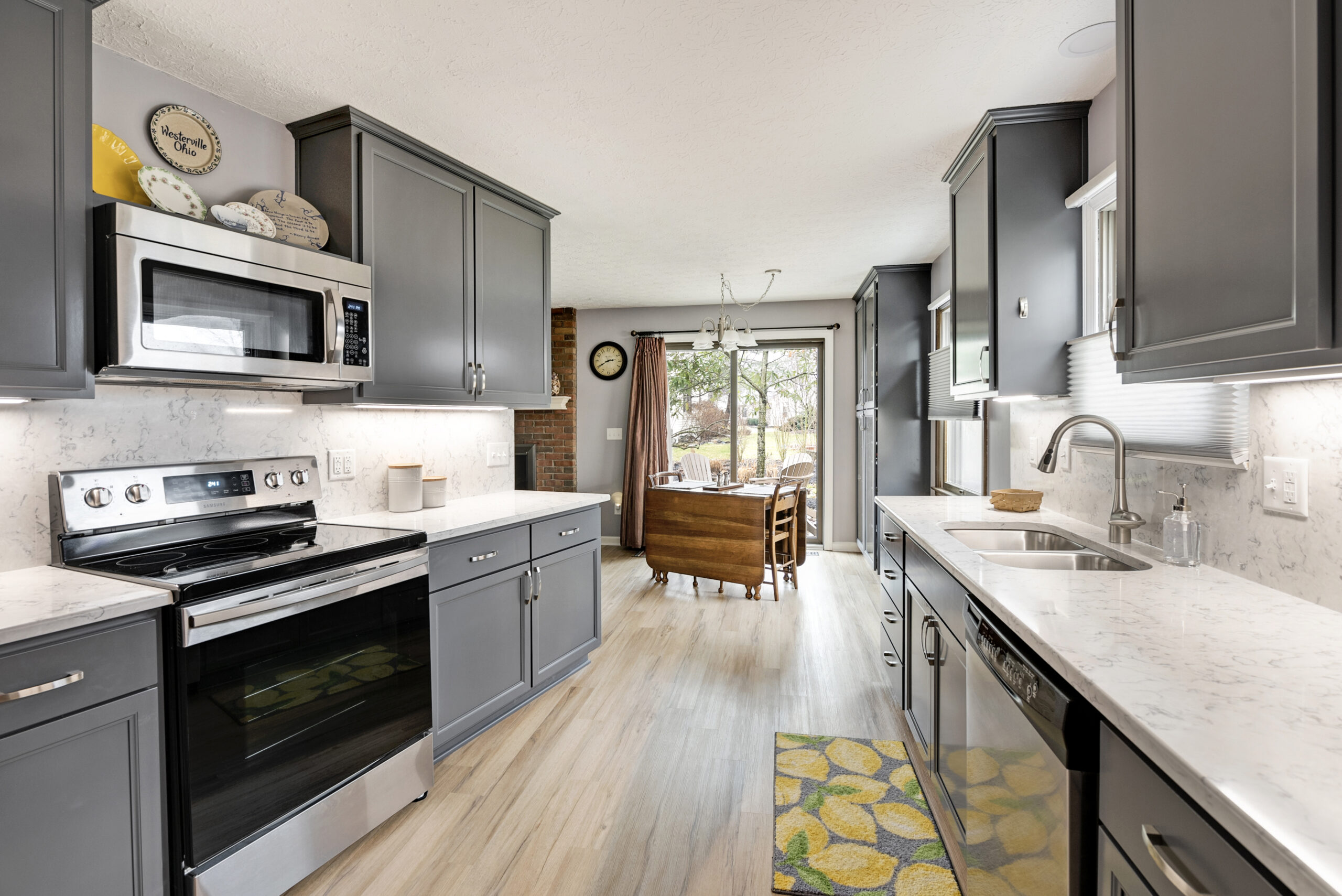 Modern kitchen with gray cabinets, stainless steel appliances, and white countertops; a dining area with large windows and a wooden table is visible in the background. Trust this kitchen remodeling company in Columbus, Ohio for stylish updates throughout your home.