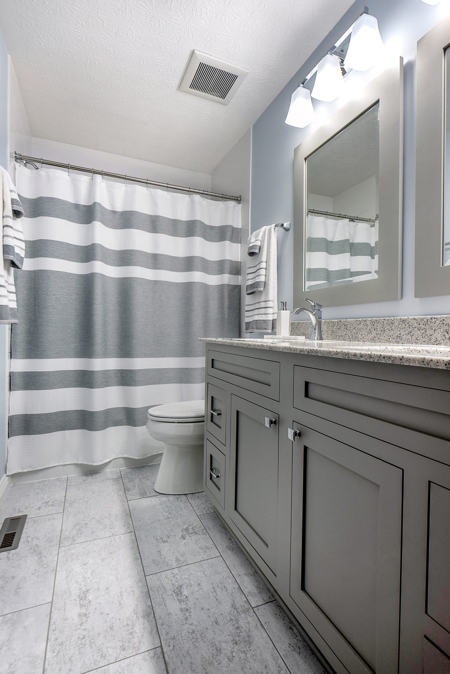 Modern bathroom with gray tile floor, light gray vanity with granite countertop, and a shower with a white and gray striped curtain—bright, clean, and neatly organized by the team behind your favorite kitchen remodel.