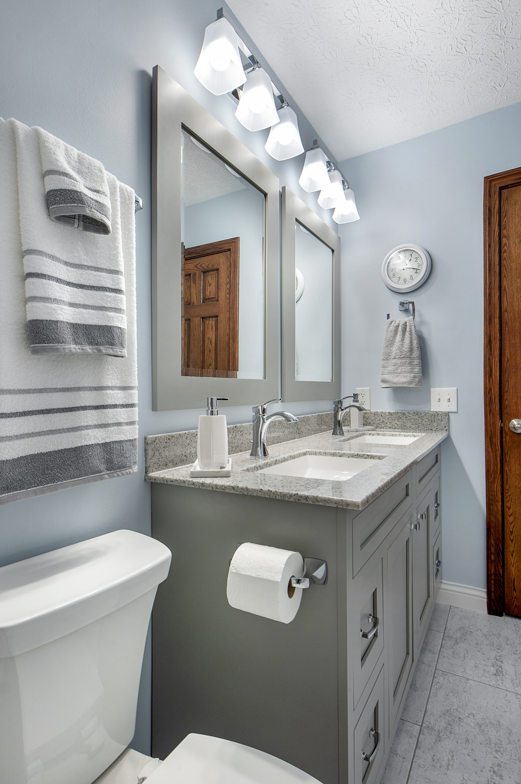 A modern bathroom with a double sink vanity, two mirrors, bright lights, a white toilet, striped towels, and light blue walls. The wooden door adds warmth to the clean decor—perfect inspiration for your bathroom renovation.