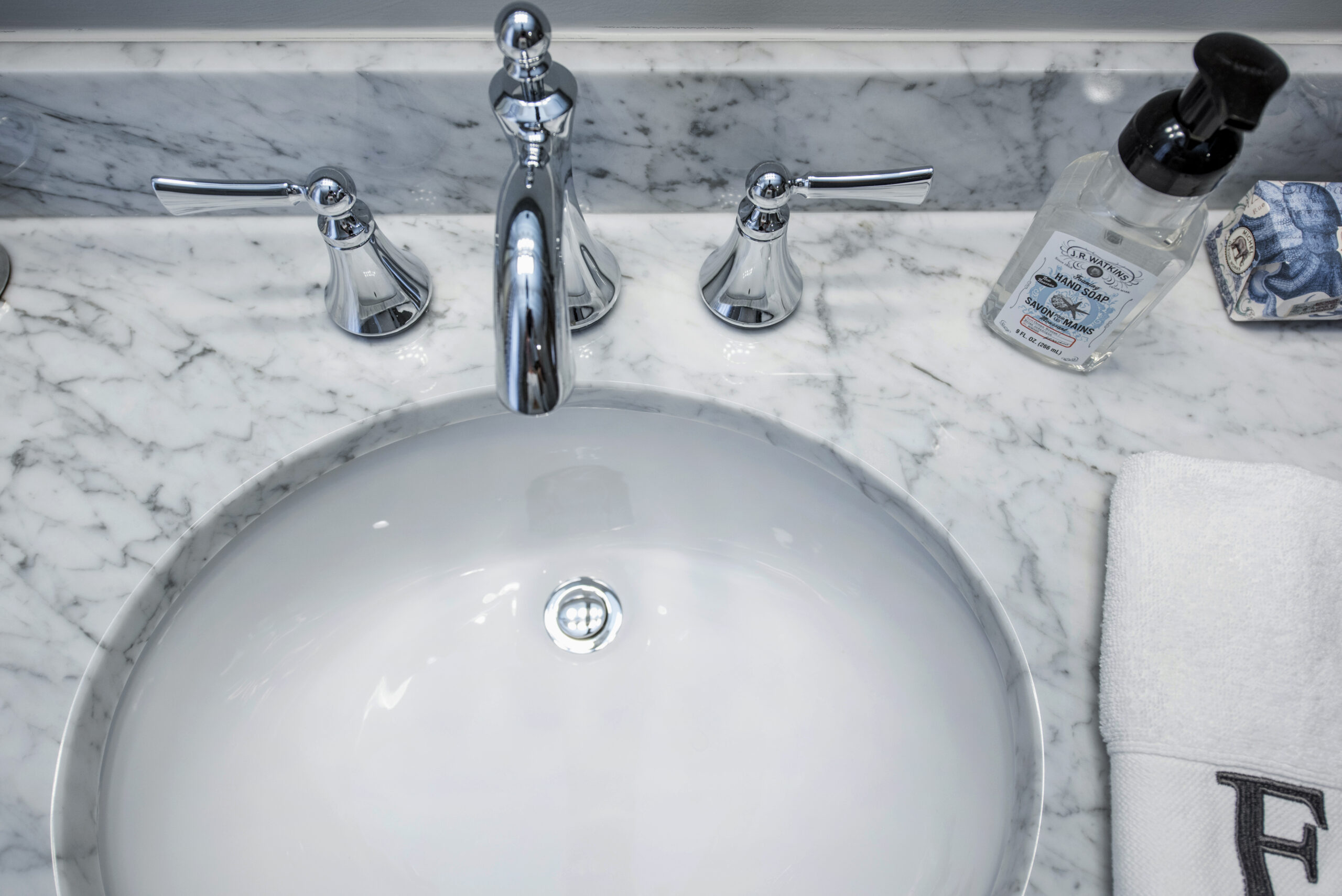 A marble bathroom countertop with a white oval sink, chrome faucet and handles, a clear soap dispenser, a patterned soap dish, and a white folded towel with a black letter “F” on it—perfect inspiration for your next bath remodel.