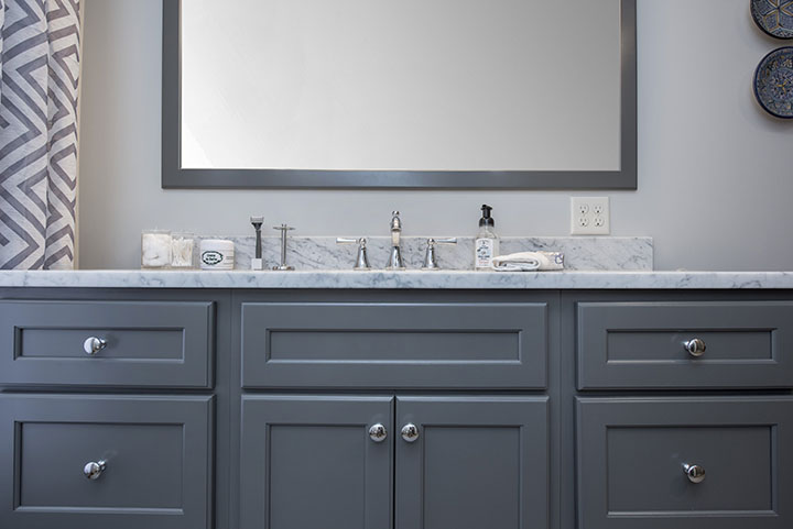 A modern bathroom vanity with grey cabinets, a marble countertop, a silver faucet, soap dispensers, and a large framed mirror above—perfect inspiration for your next bath remodel. The background features a patterned shower curtain and decorative wall plates.