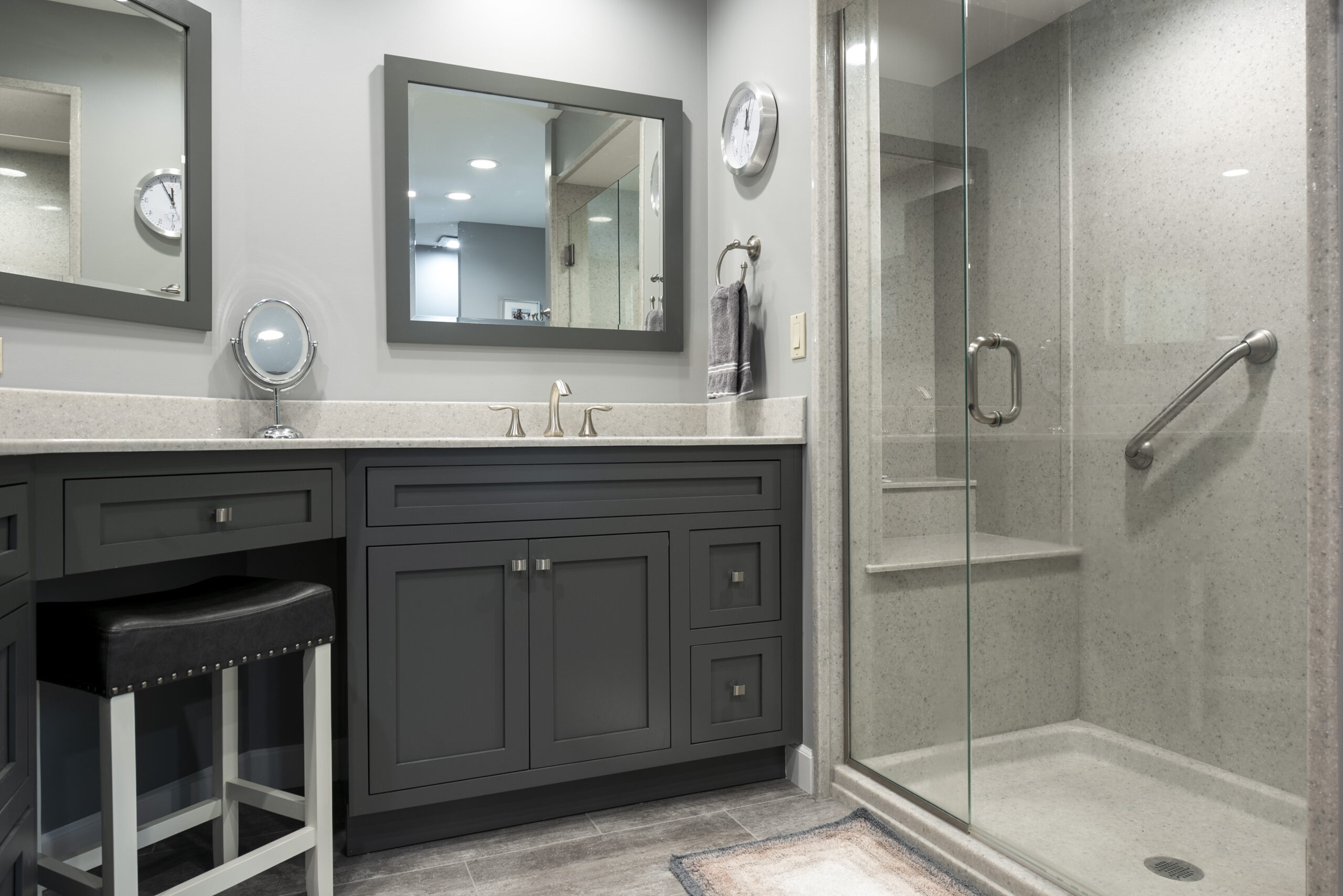 Modern bathroom by a leading bathroom remodeling company in Columbus, Ohio, featuring gray cabinets, wide vanity with mirror, small stool, and a walk-in glass shower with built-in bench and metal grab bar. A wall clock and neat countertop complete the clean look.