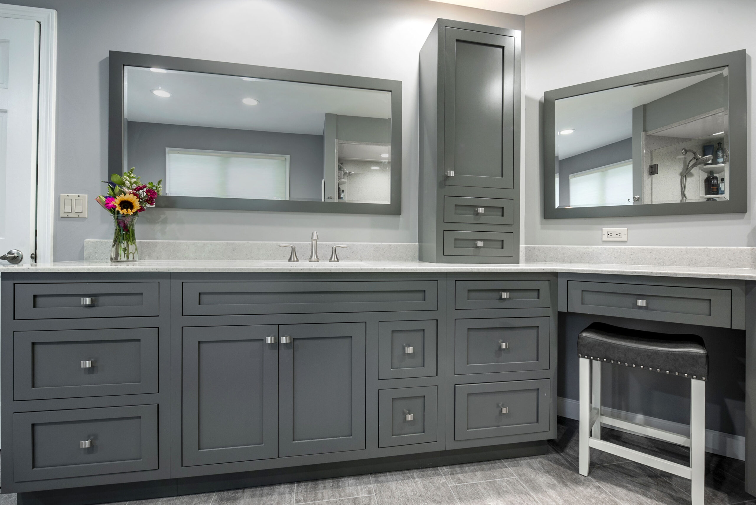 Modern bathroom with gray cabinetry, two large mirrors, silver fixtures, a flower vase on the left counter, and a small vanity area with a black cushioned stool—perfect inspiration for your next bath remodel.