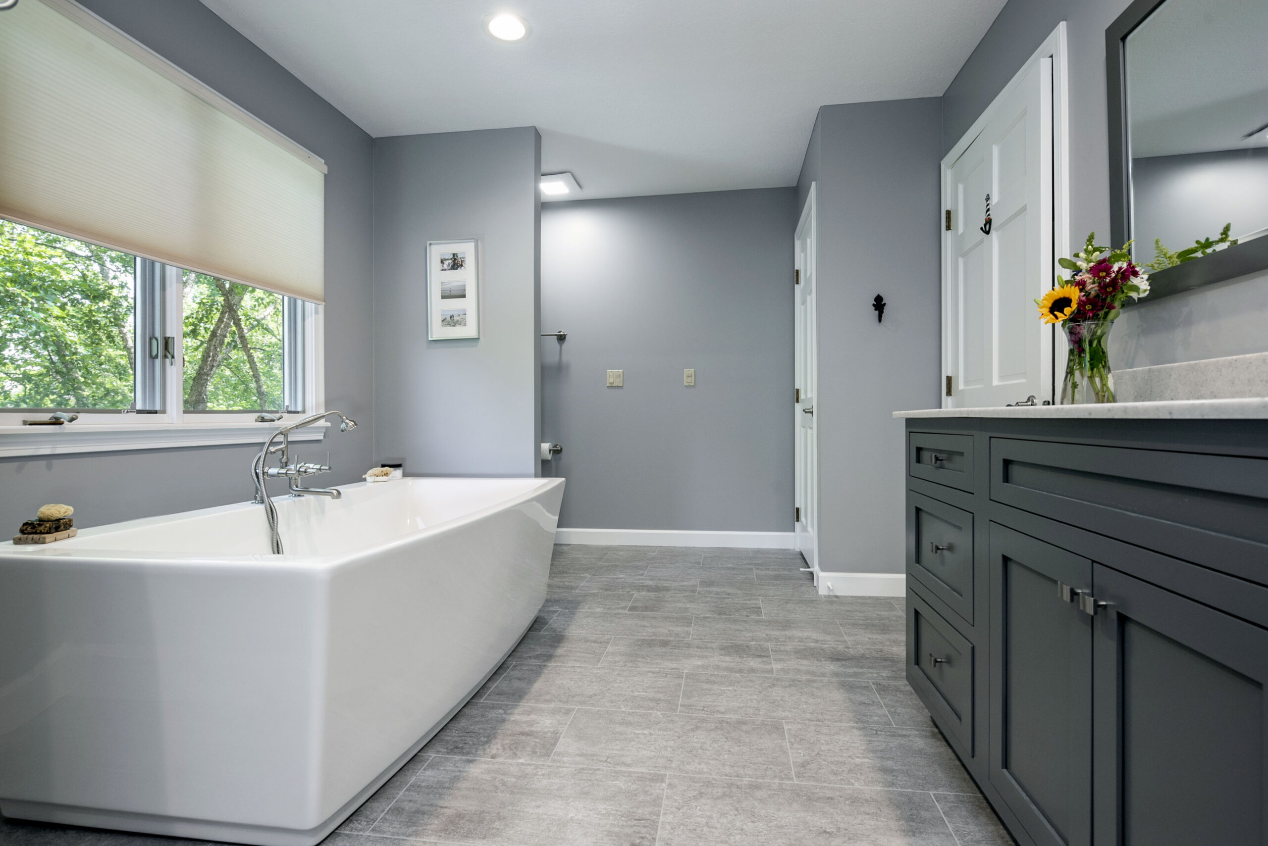 Modern bathroom with large white soaking tub by a window, gray walls, tiled floor, and gray vanity with a mirror and vase of sunflowers. Bright natural light fills the space—beautiful work by a leading bathroom remodeling company in Columbus, Ohio.