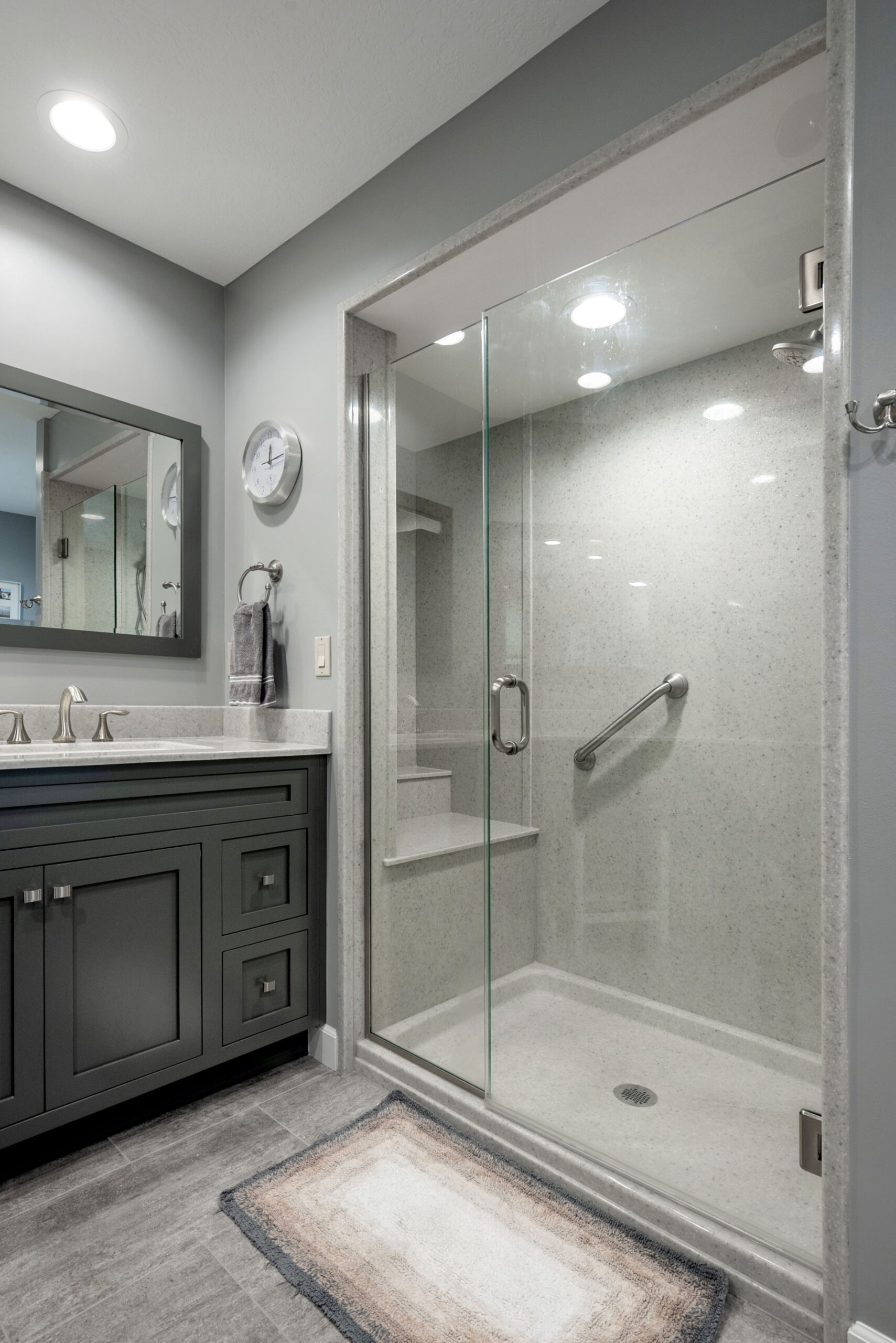 Modern bathroom with gray walls, a glass-enclosed shower featuring a grab bar and built-in bench, a dark vanity with a sink and mirror, and a rectangular rug on the gray tile floor by a top bathroom remodeling company in Columbus, Ohio.