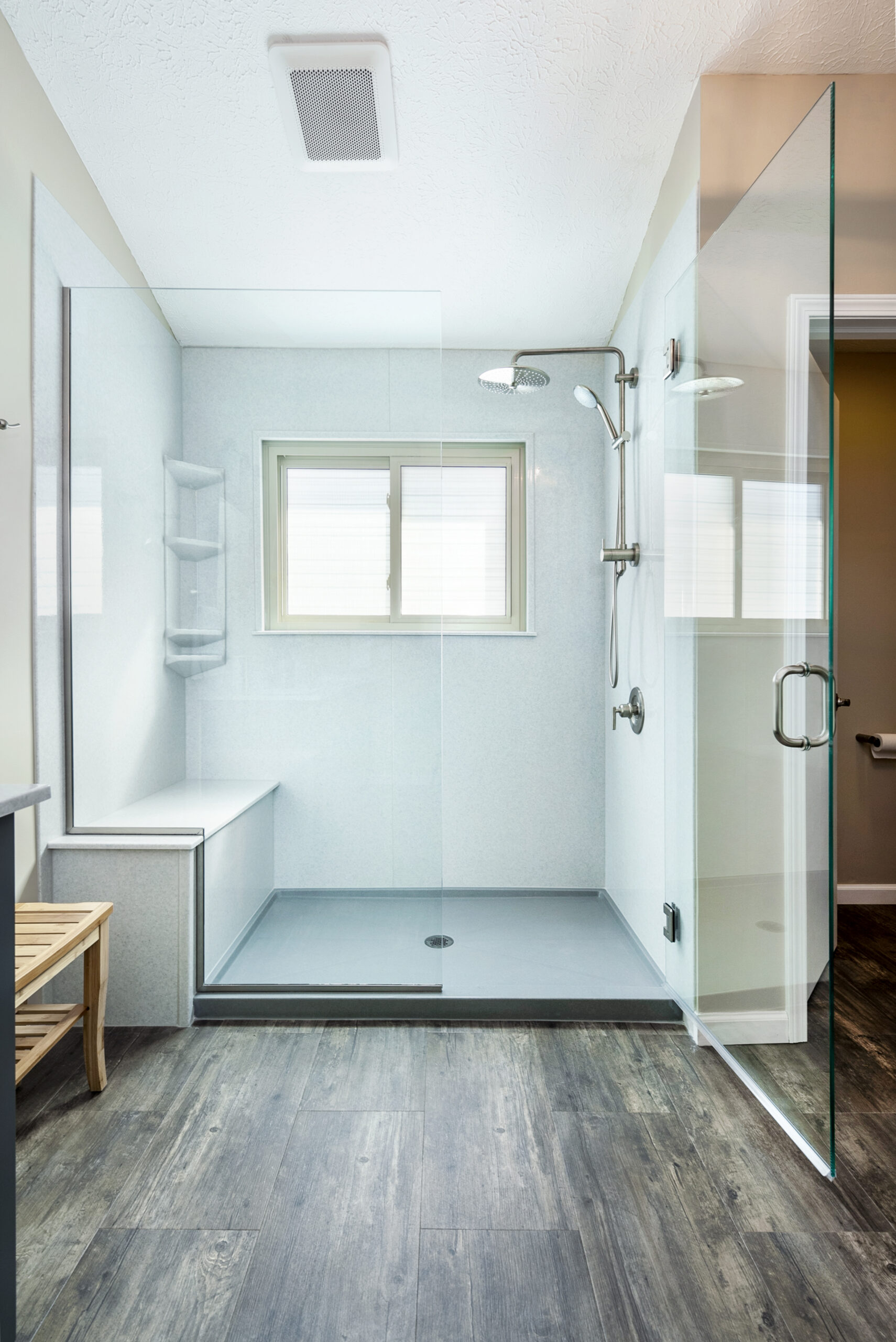Modern walk-in shower with glass doors, a built-in bench, wall shelves, and a rainfall showerhead—perfect for your next bathroom renovation. The shower features light walls, a frosted window, dark wood flooring, and a wooden bench just outside.