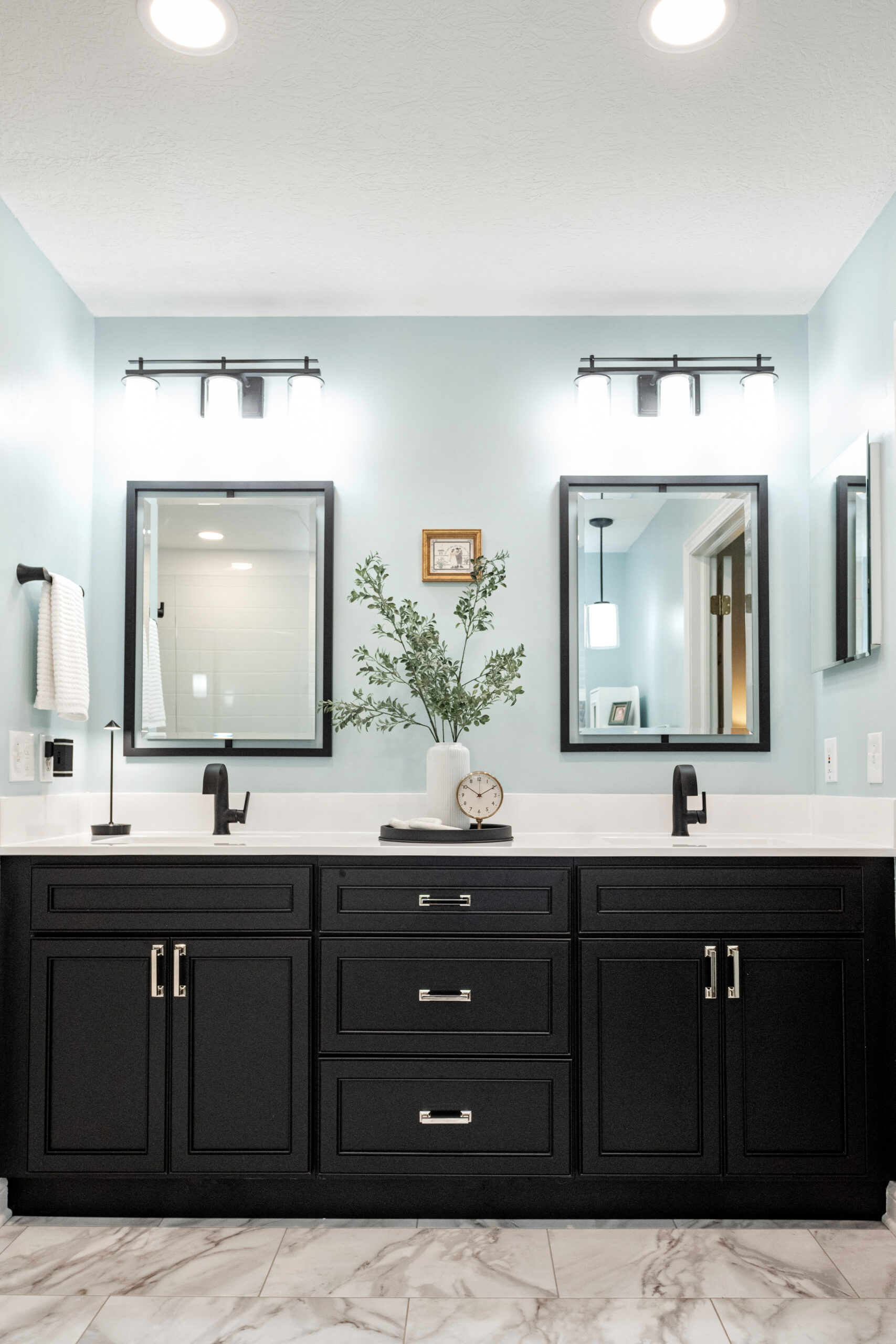 Modern bathroom by a leading bathroom remodeling company in Columbus, Ohio, featuring a black double-sink vanity, white countertop, two lighted mirrors, small plant, clock, towel on the wall, light blue walls, and marble tile floor.