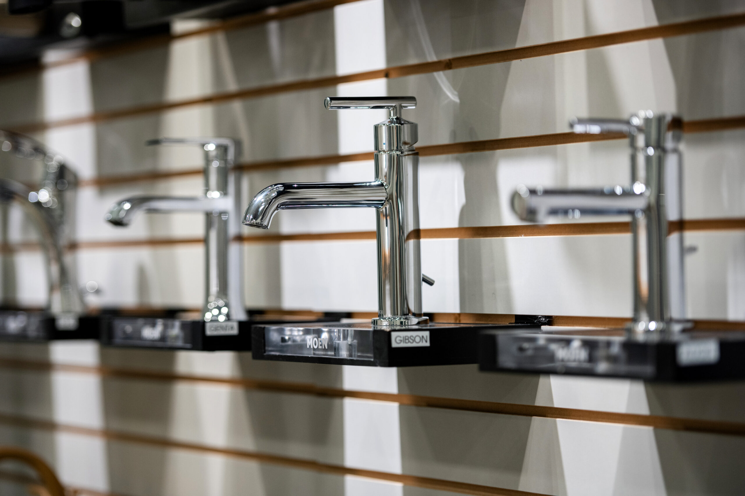 Several modern chrome bathroom faucets are displayed on black stands against a wall with horizontal wooden slats—perfect inspiration for your next bathroom renovation or kitchen remodel.