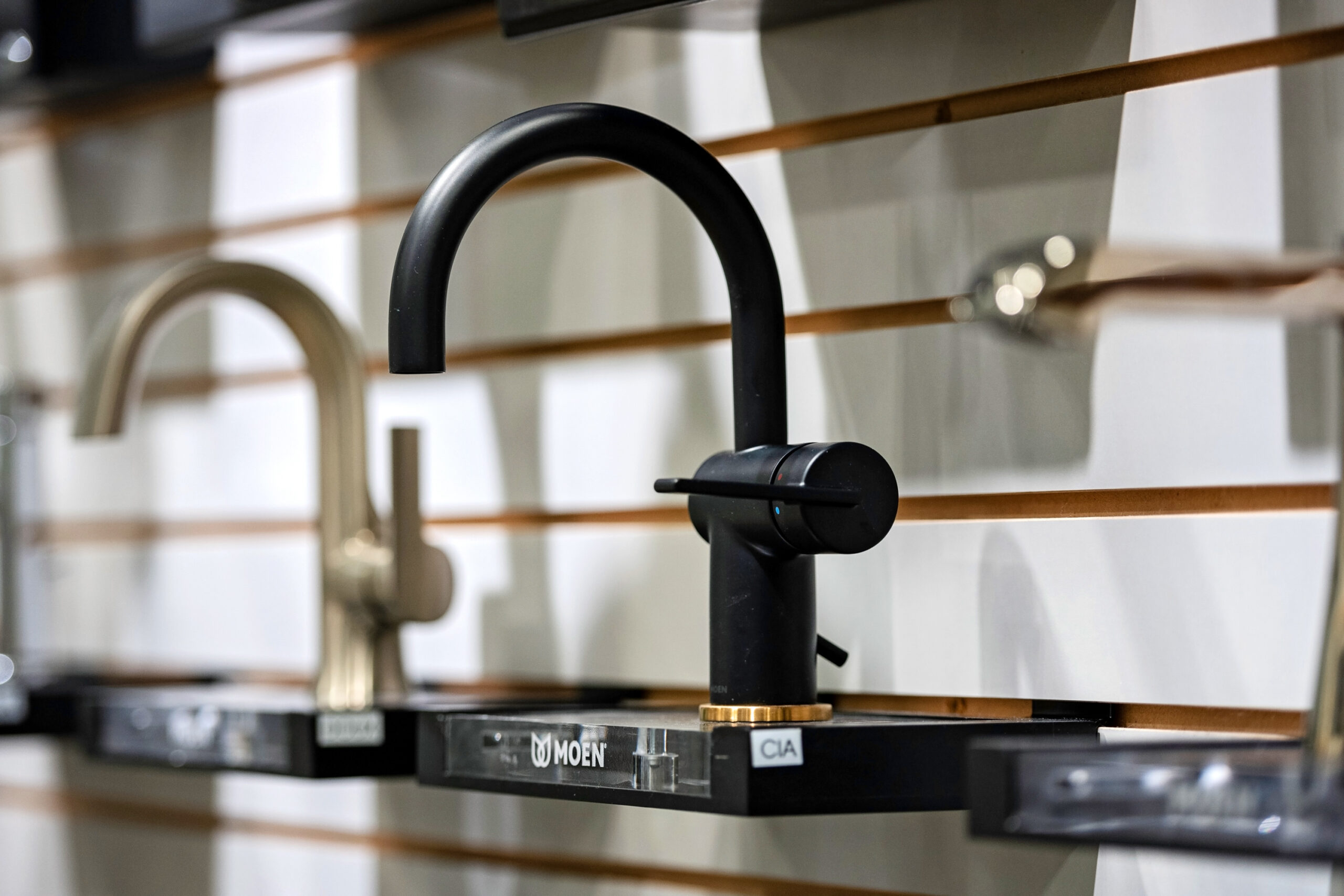 A display of modern kitchen faucets, perfect for a kitchen remodel, features a matte black faucet with a curved spout and single lever handle, mounted on a wall alongside other sleek metallic models.