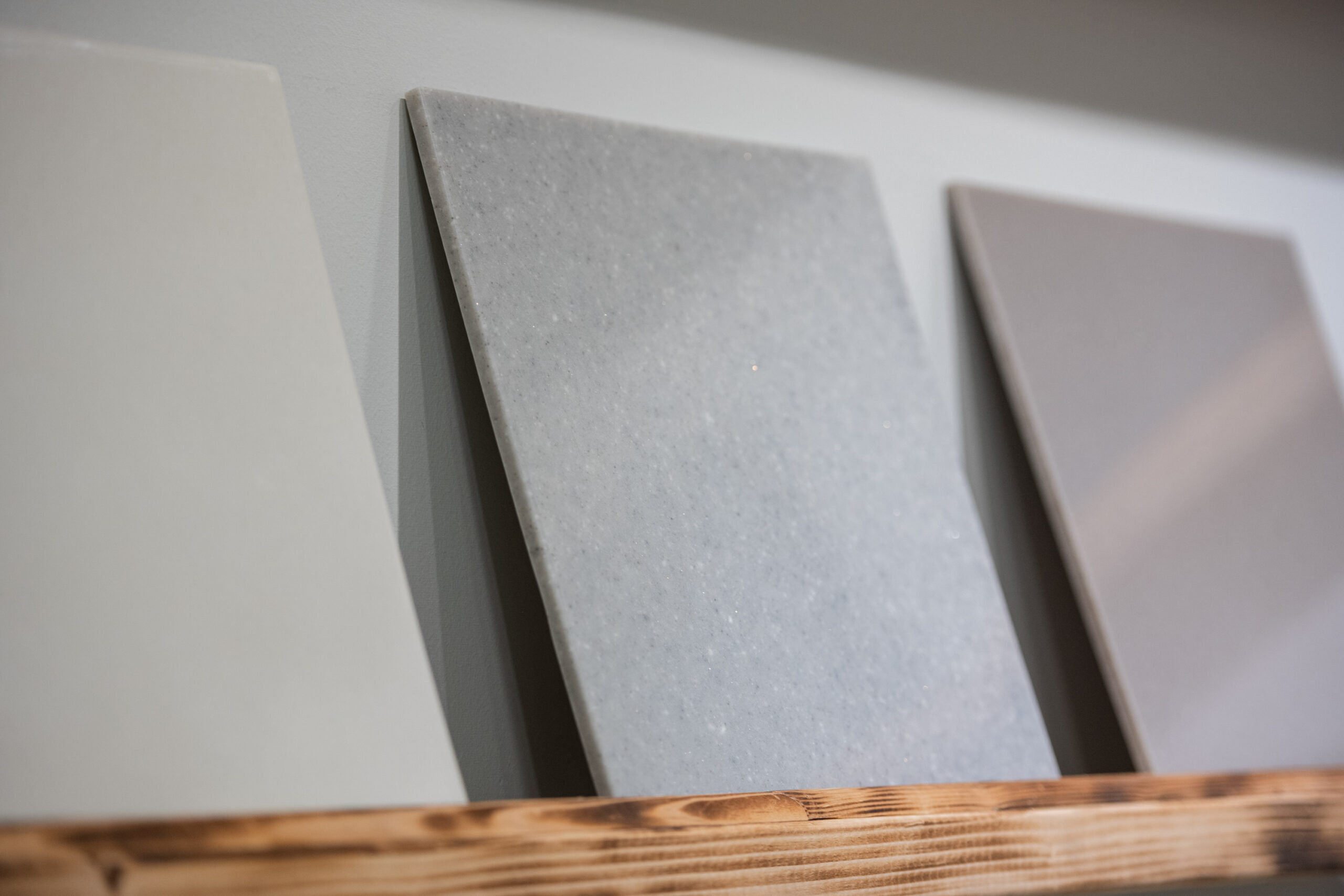 Three large rectangular samples of gray and beige solid surface material are displayed upright on a wooden shelf—perfect inspiration for your next kitchen renovation or bathroom remodeling project in Columbus, Ohio.