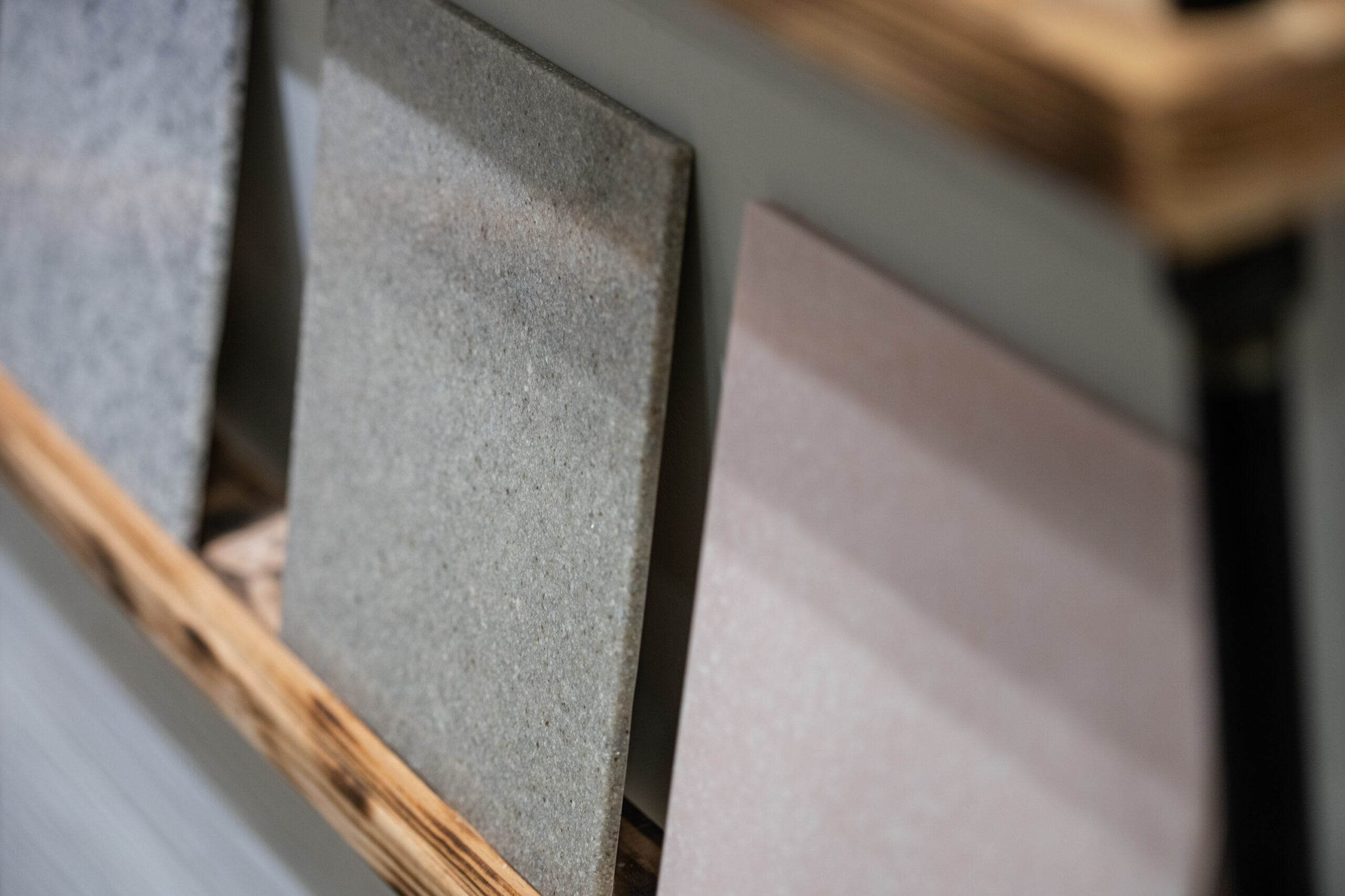 Close-up of three rectangular ceramic tiles in neutral tones, displayed upright on a wooden rack against a gray background—an inspiring choice for your next kitchen remodel.