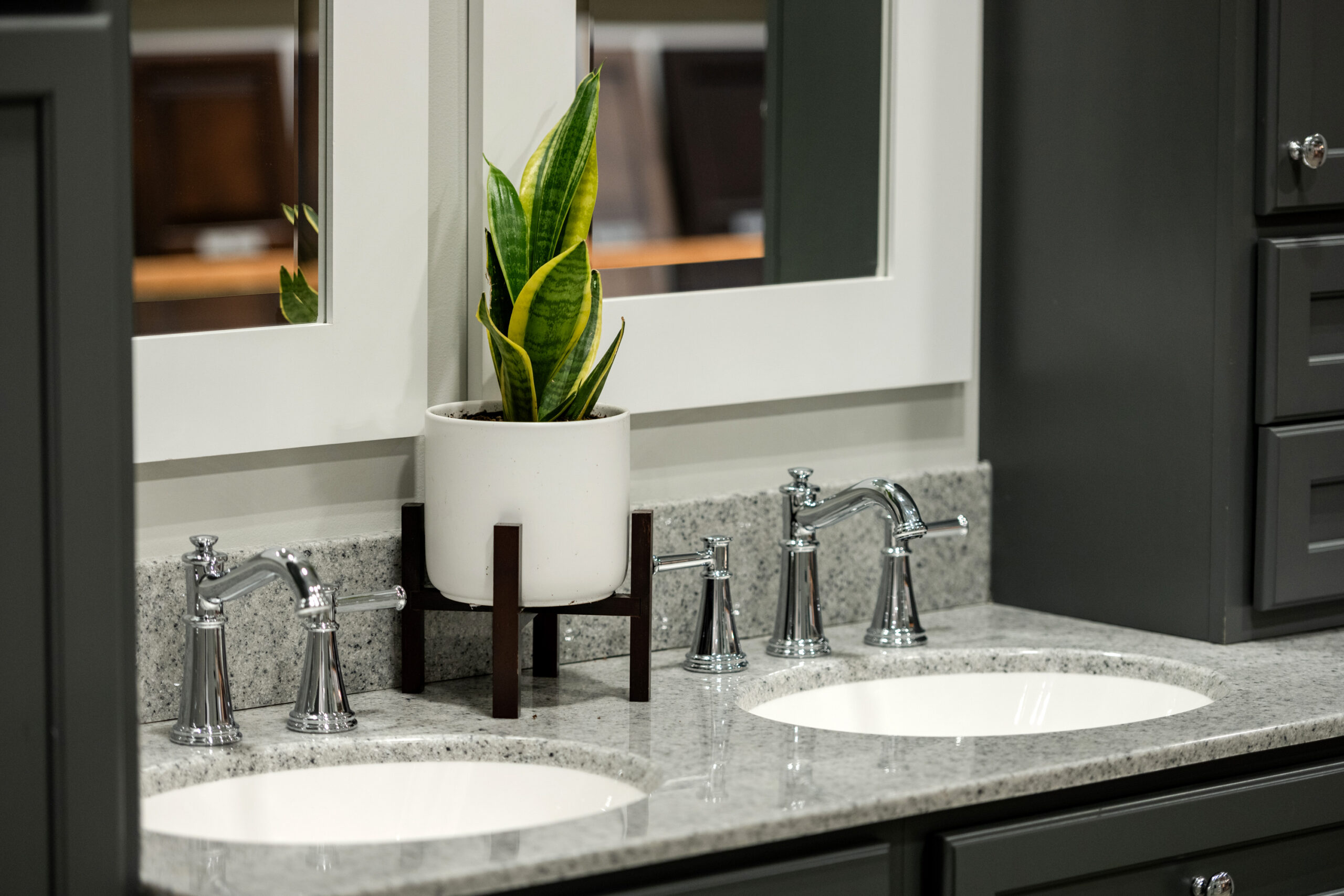 A modern bathroom vanity with two sinks, chrome faucets, and a granite countertop features a potted snake plant on a wooden stand between two mirrors—perfect inspiration from a top bath remodel or bathroom remodeling company in Columbus, Ohio.