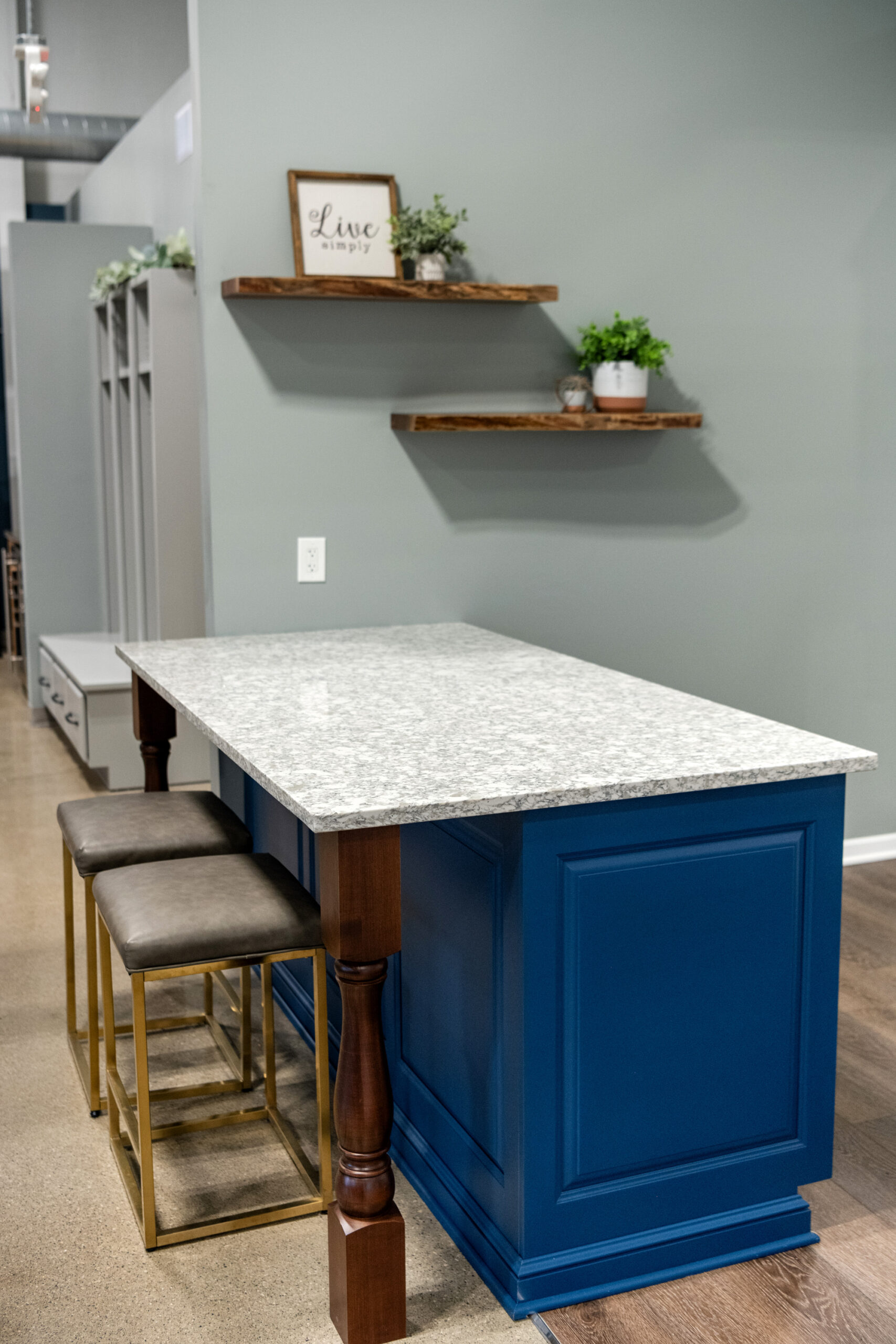 A kitchen remodel features a blue island with a light granite countertop, paired with two stools. Wooden shelves adorned with small plants and decor are mounted on a gray wall—perfect inspiration from a top kitchen remodeling company in Columbus, Ohio.