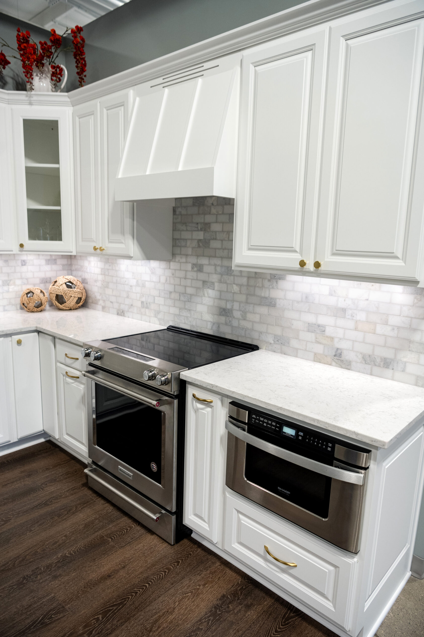 A modern kitchen renovation with white cabinets, marble backsplash, stainless steel oven and microwave, and light countertops. Decorative items sit on the counters, complemented by a dark wood floor—crafted by a top kitchen remodeling company Columbus Ohio.