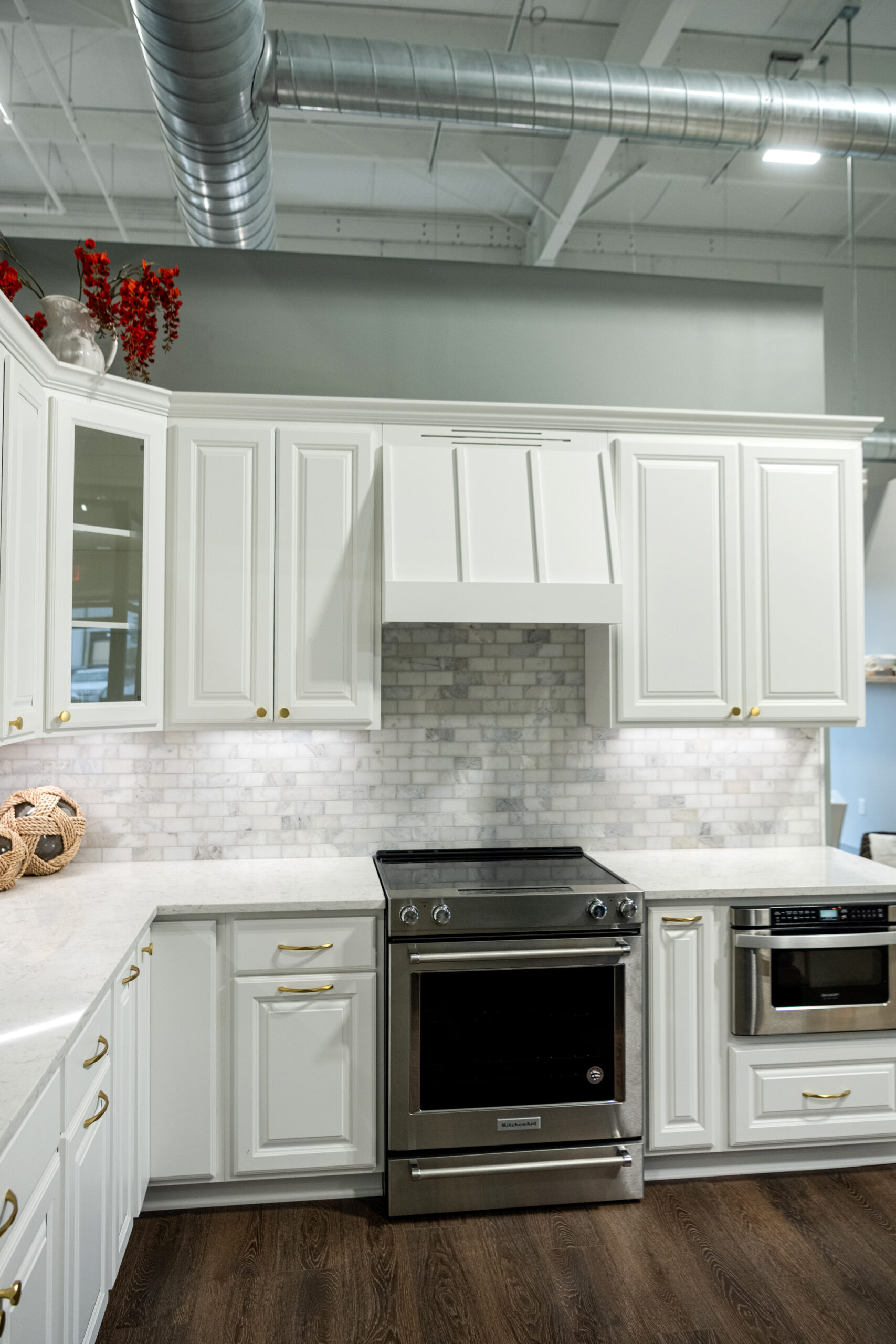 Modern kitchen with white cabinets, stainless steel stove and microwave, marble backsplash, and dark wood floors. Red flowers and a decorative bowl sit on the counters. Exposed ductwork highlights the style by this kitchen remodeling company Columbus Ohio.