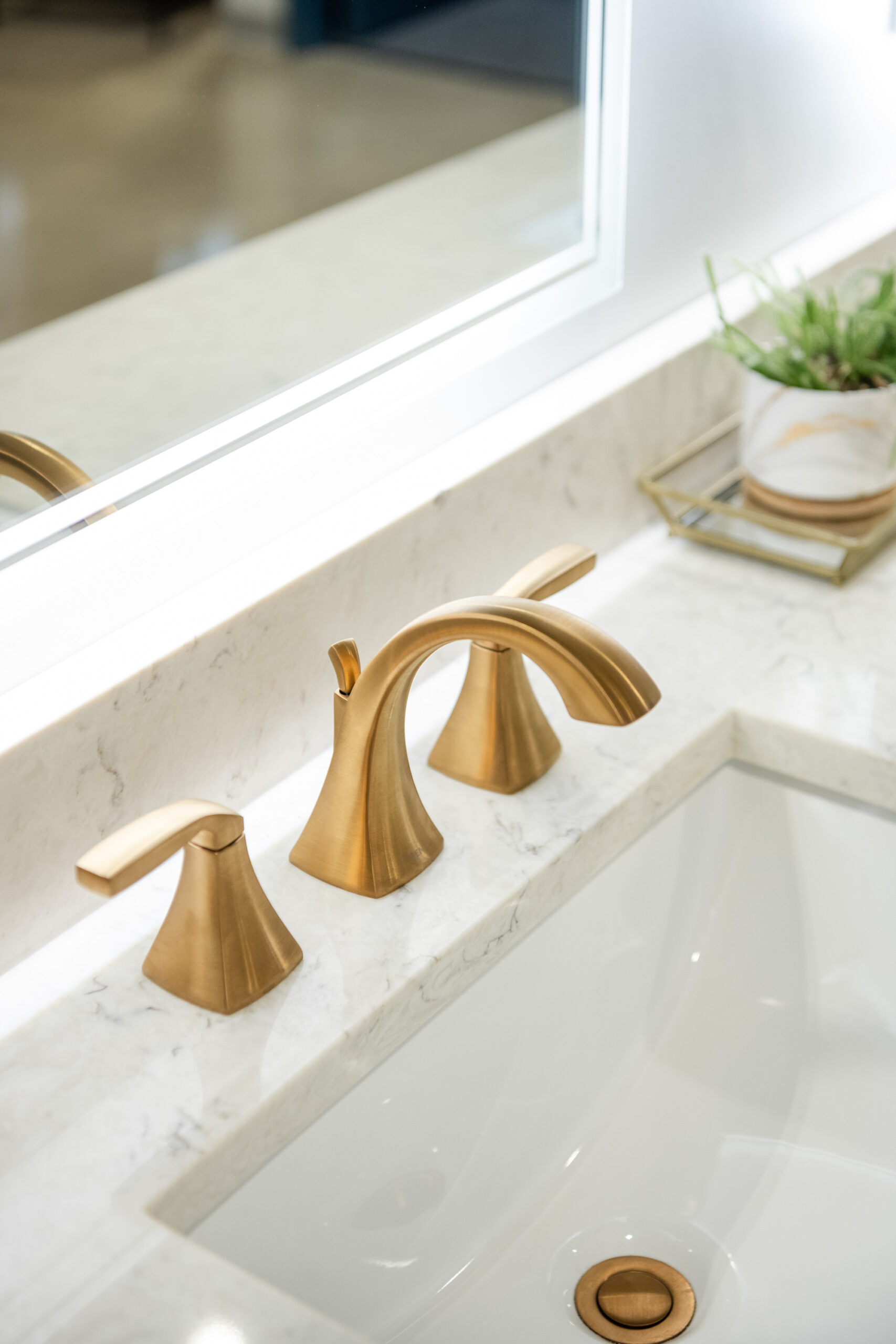 Close-up of a modern bathroom sink with a gold faucet set on a white marble countertop, styled by a top bathroom remodeling company in Columbus, Ohio. A small green potted plant and a rectangular mirror complete the elegant look.