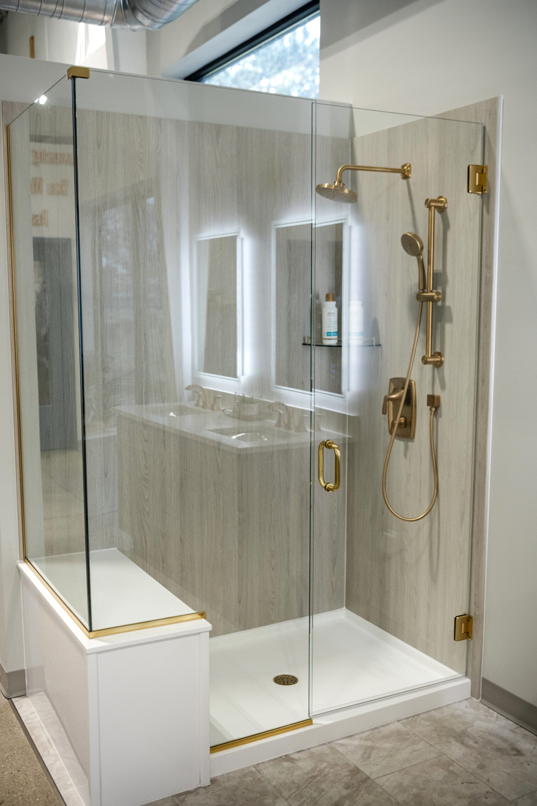 A modern glass shower with gold fixtures, including a rainfall showerhead and handheld sprayer, sits next to a wall-mounted vanity with two lighted mirrors in a bright bath remodel showroom by a leading bathroom remodeling company in Columbus, Ohio.