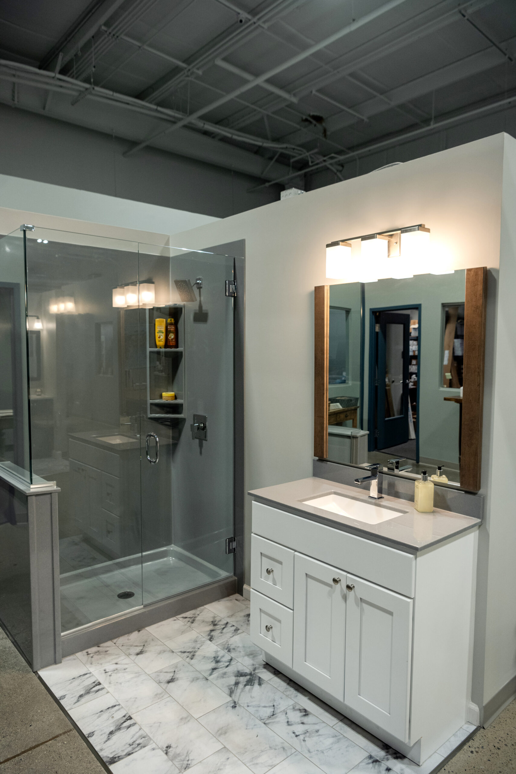 Modern bathroom display featuring a glass-enclosed shower with shelves, a white vanity with drawers, a rectangular sink, and marble-patterned floor tiles—perfect inspiration for your next bathroom renovation under bright lighting.