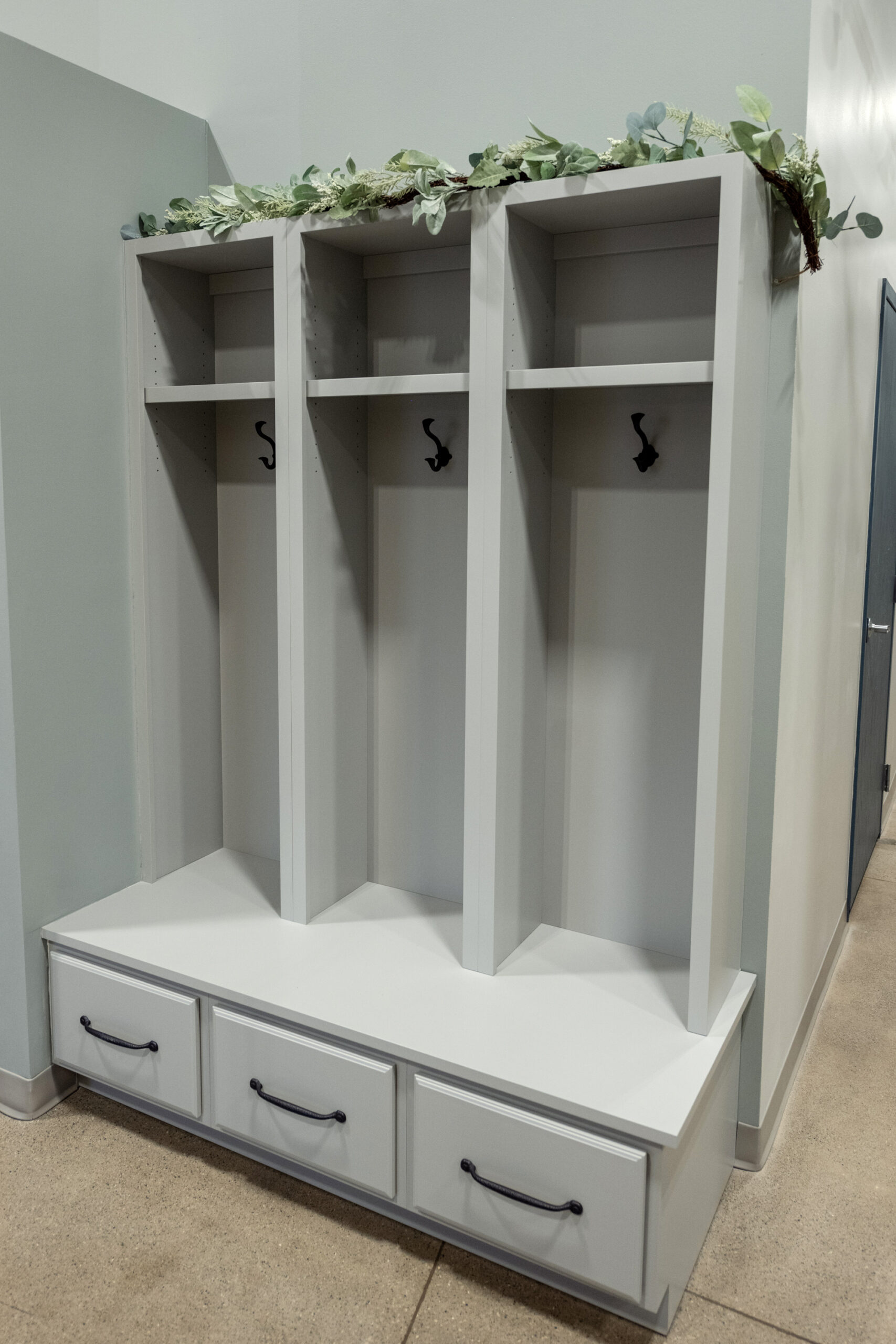 A white built-in cubby storage unit with three open sections, each with a hook and shelf, sits against a wall—perfect for your next kitchen renovation. Three drawers are below, green leafy decor lines the top, and tan floors complement light walls.