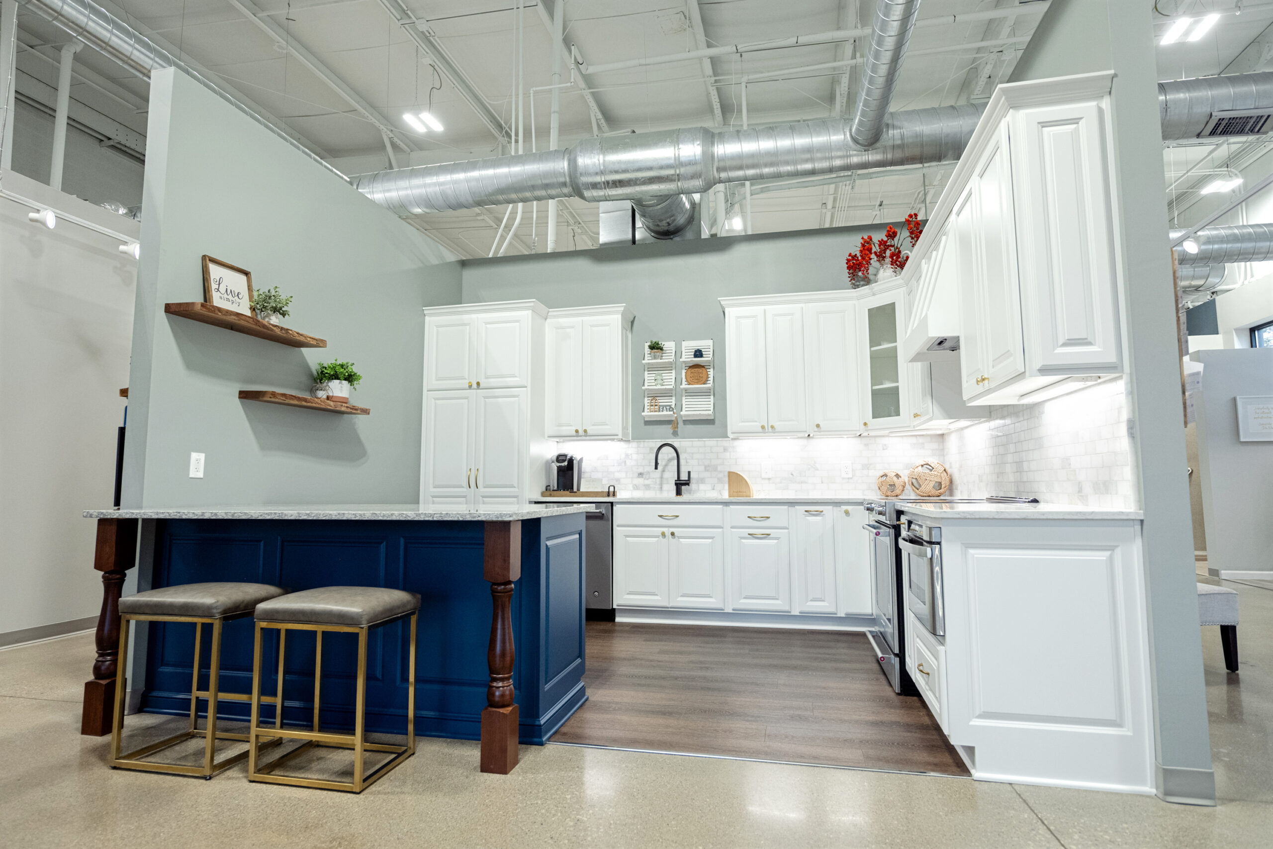 Modern kitchen renovation with white cabinets, stainless steel appliances, and a blue island with two upholstered stools. Open shelving with plants and decor, tiled backsplash, and exposed ductwork on the ceiling.